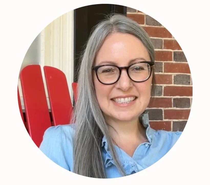 A woman with long gray hair and glasses, smiling, wearing a light blue shirt, sitting outdoors with a red Adirondack chair and a brick wall in the background.