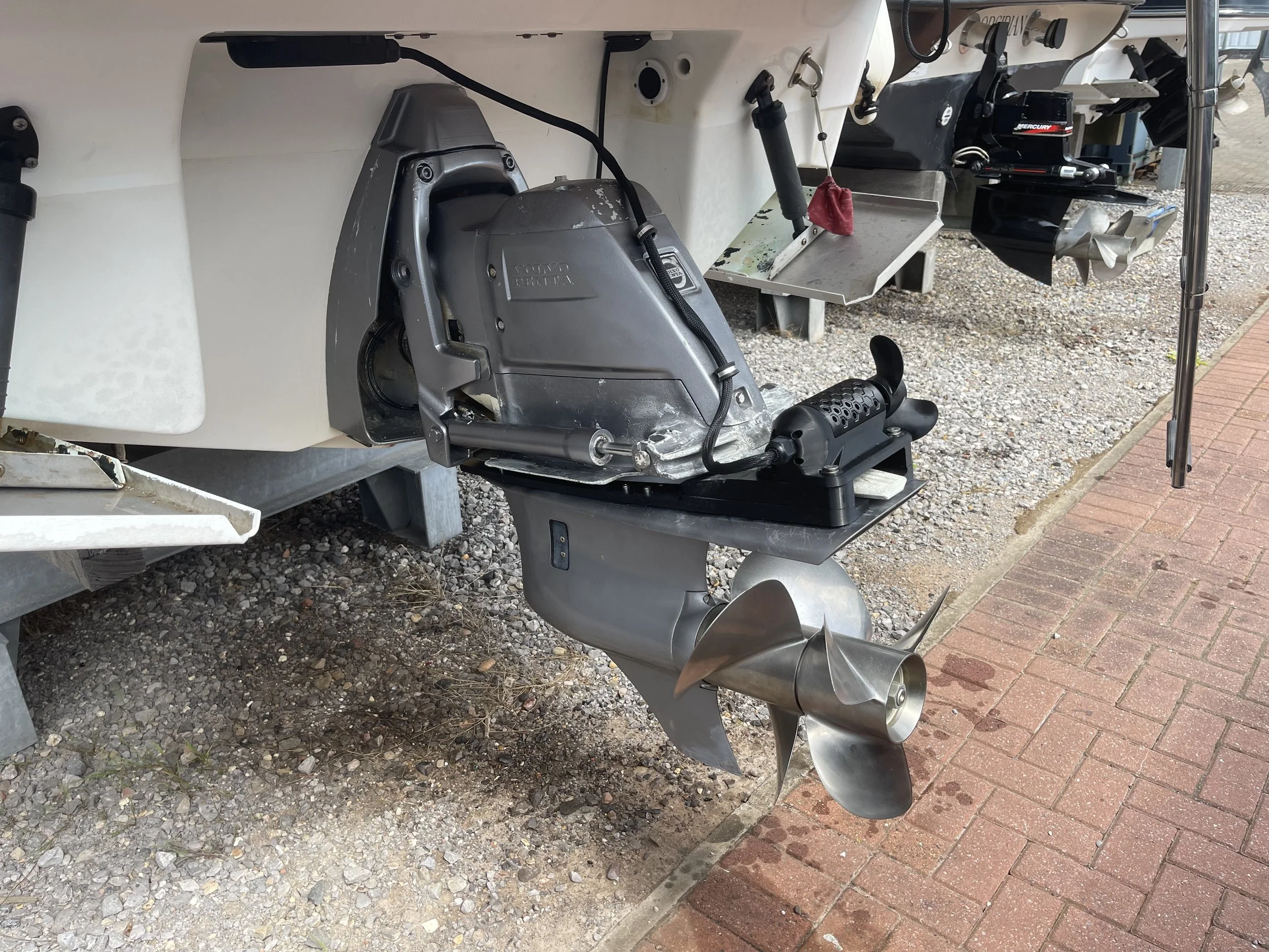 Close-up of the GTD Marine SidePod stern thruster and outboard propeller of a boat, with some black and silver components and a red cloth tied to a rod, on gravel ground and brick pavement.