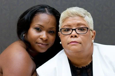 Two women posing closely, one with dark hair and the other with short blonde hair and glasses, both with neutral expressions.