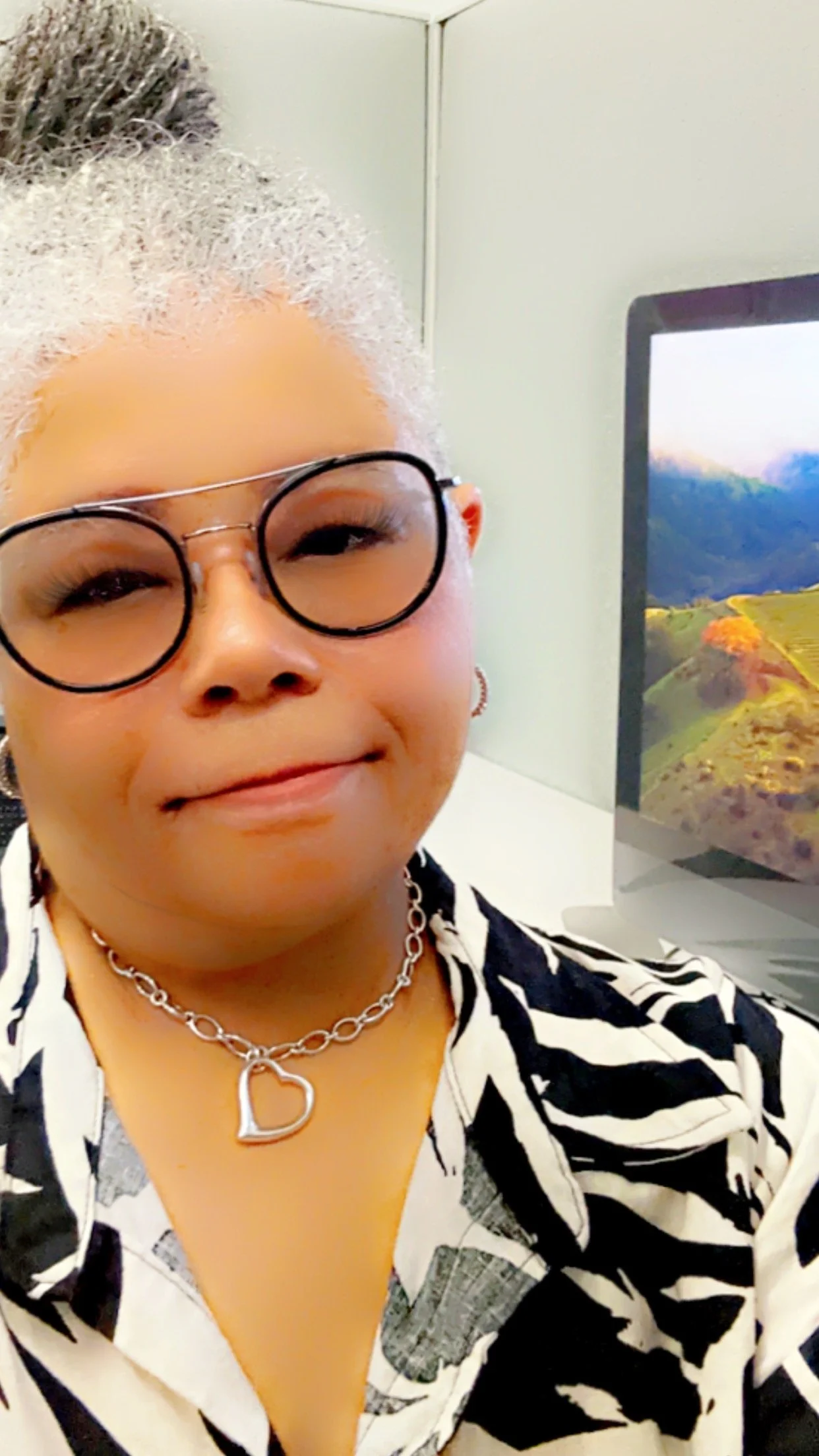 Person wearing glasses and a patterned shirt in front of a computer with landscape wallpaper.