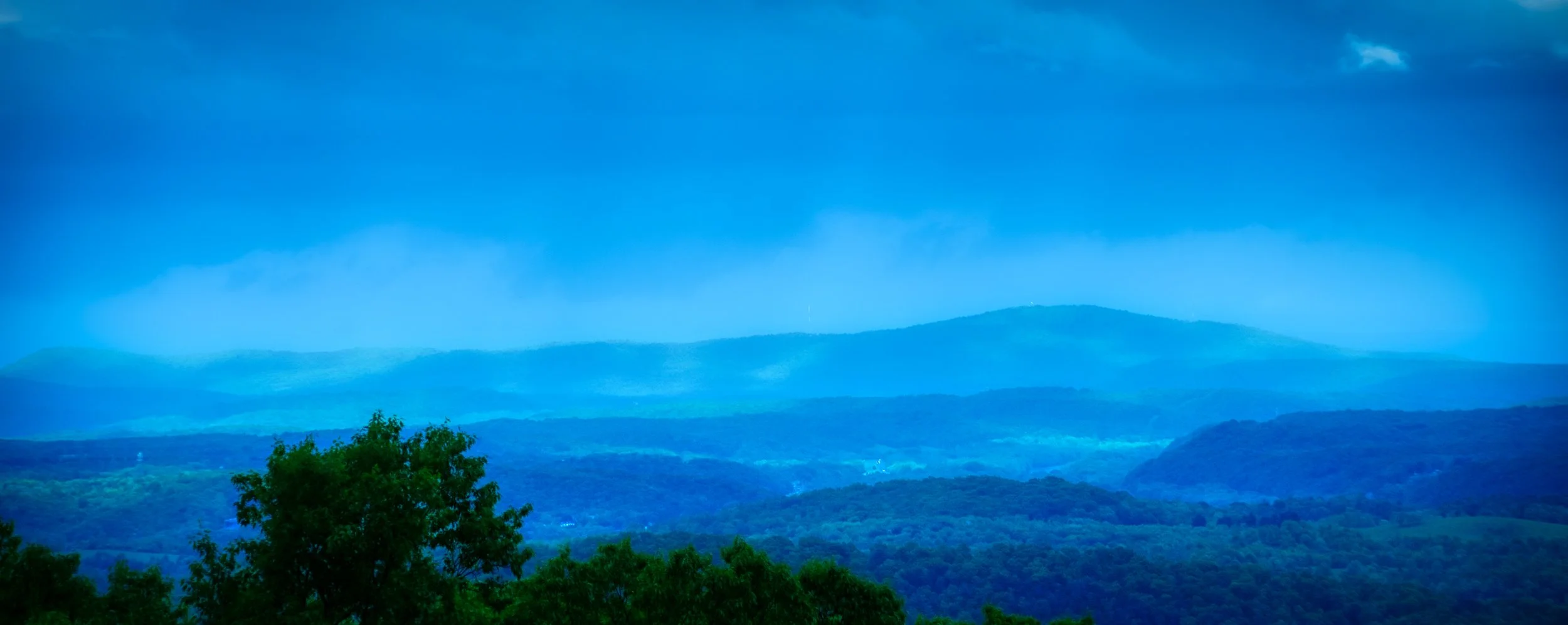 Scenic view of rolling blue hills and lush green trees under a cloudy sky.