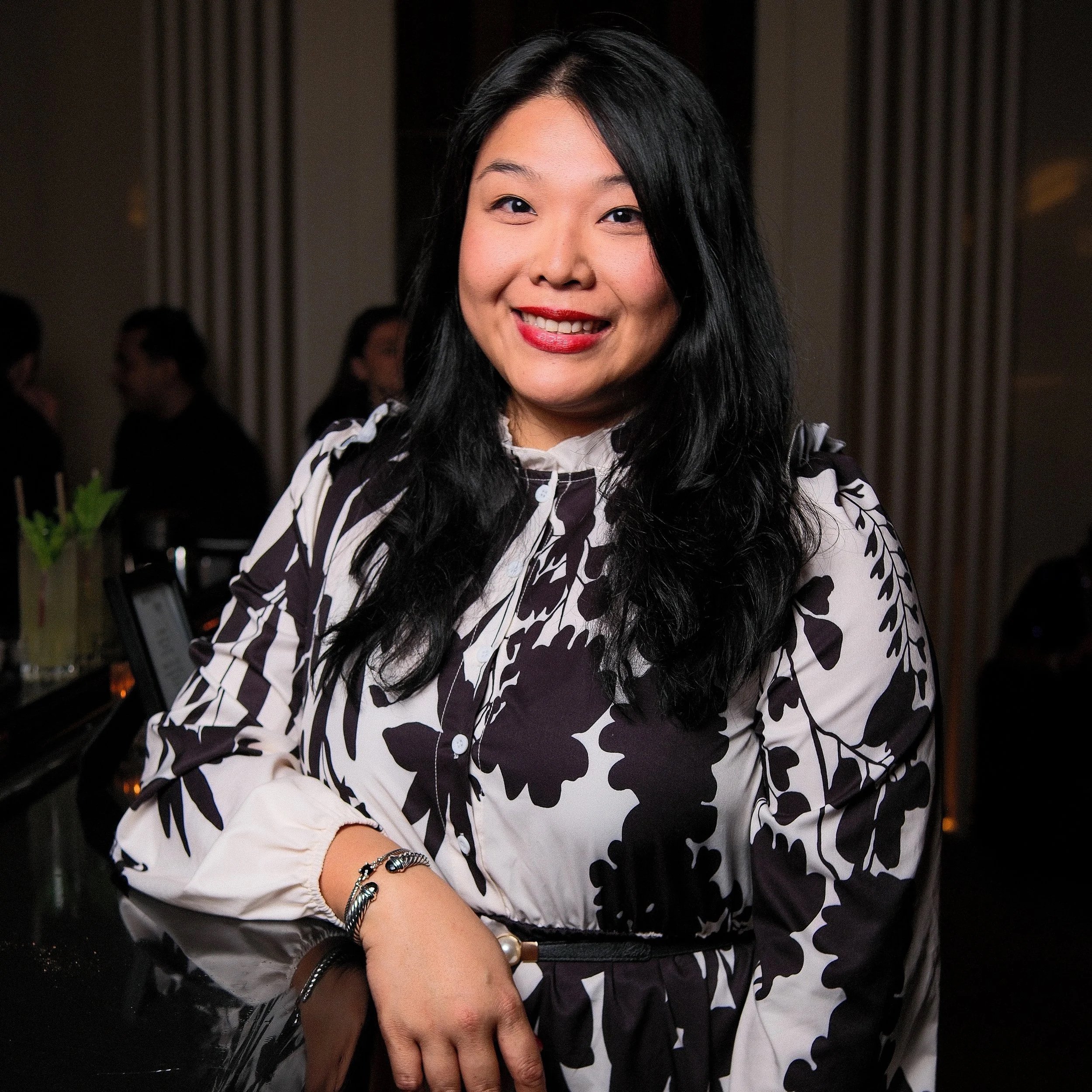 A woman with long dark hair smiling indoors at a social gathering, wearing a black and white floral patterned blouse and bracelets.