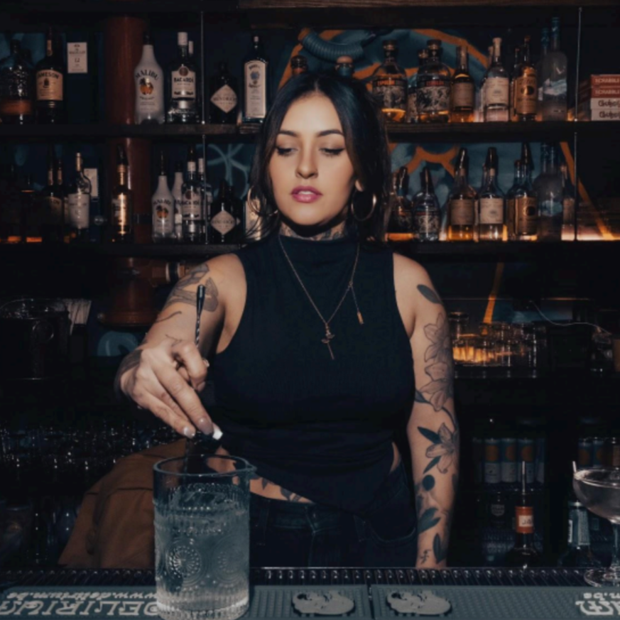A female bartender with dark hair, tattoos, and hoop earrings preparing a drink at a bar with various liquor bottles on shelves behind her.