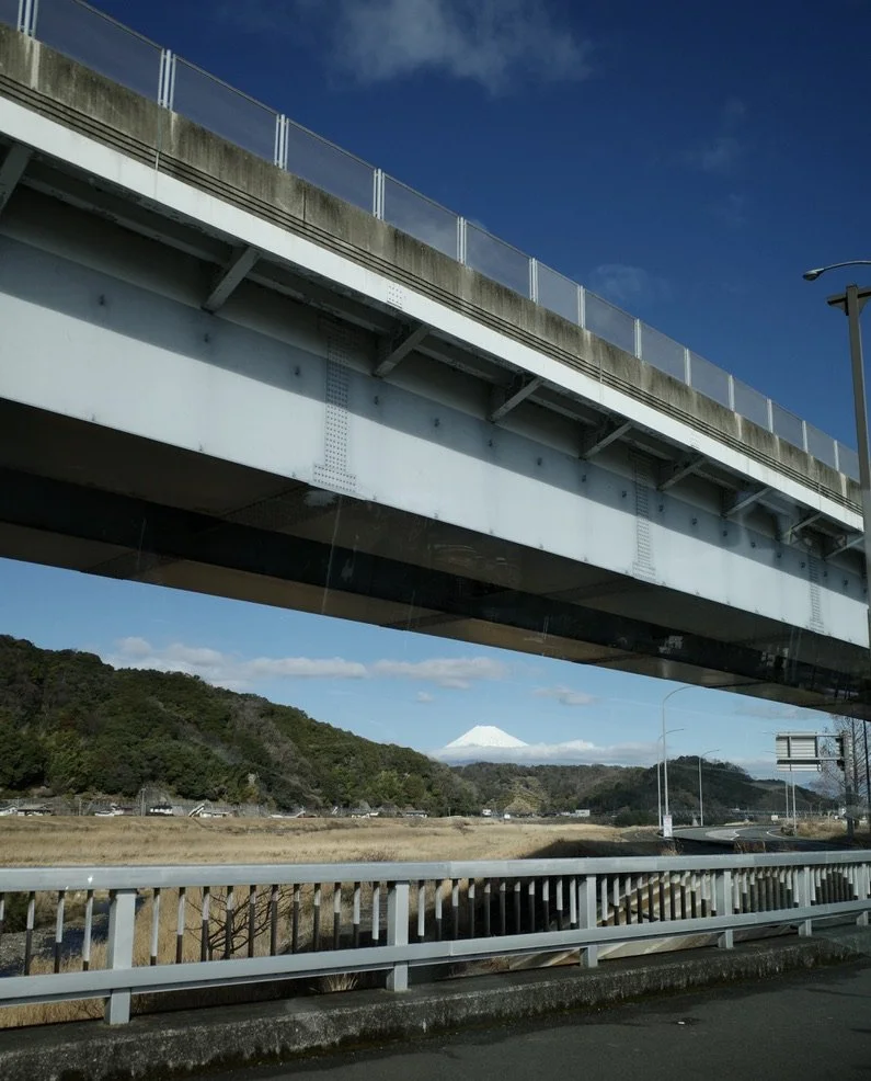 Snow‑capped mountain seen beneath an elevated highway, with grass and hills in the foreground.