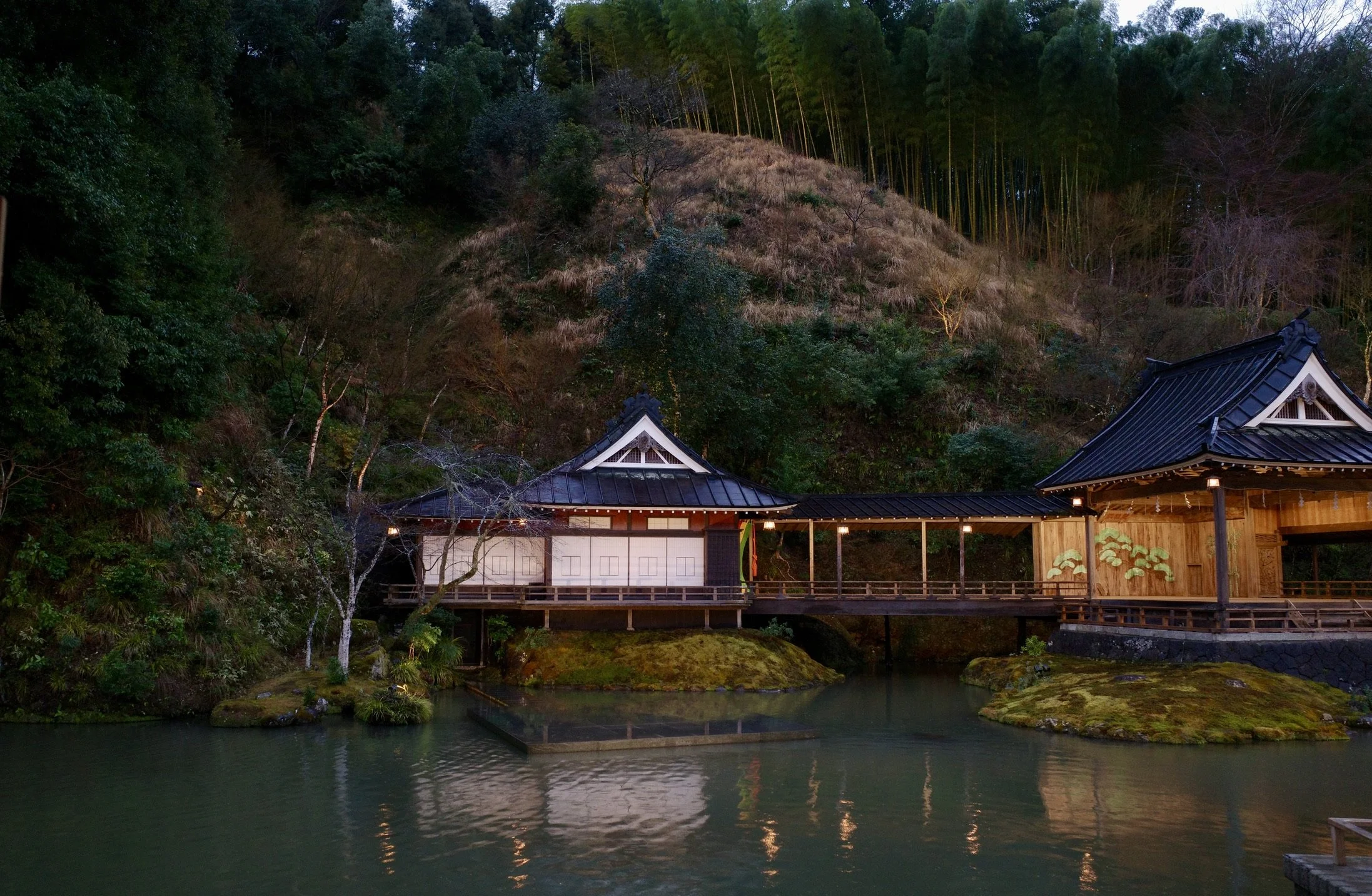 Two wooden buildings with tiled roofs connected by a covered bridge over a pond, surrounded by greenery.