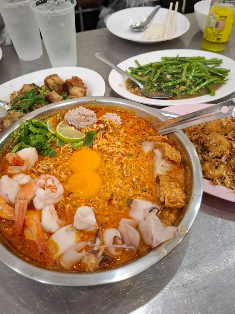 Large bowl of noodle soup topped with shrimp, squid, greens, lime, and raw egg yolks, surrounded by side dishes.