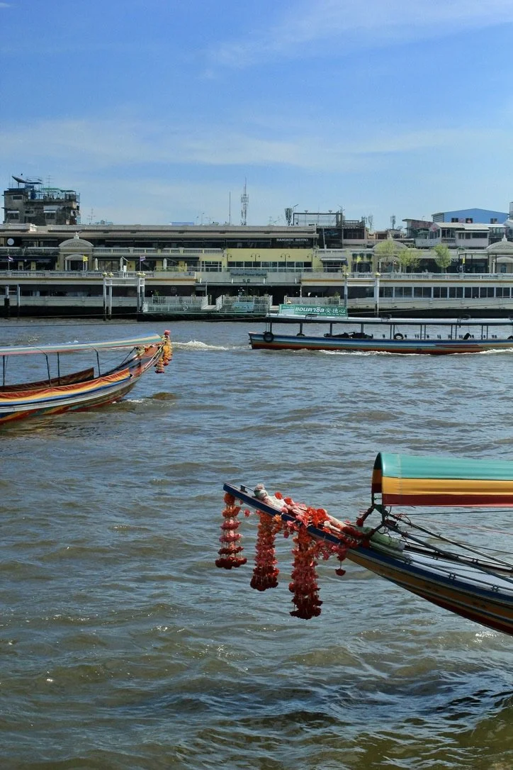 Long‑tail boats with colorful painted hulls and red flower garlands move along a river, with multi‑story buildings and smaller riverside structures visible on the opposite bank under a clear blue sky.