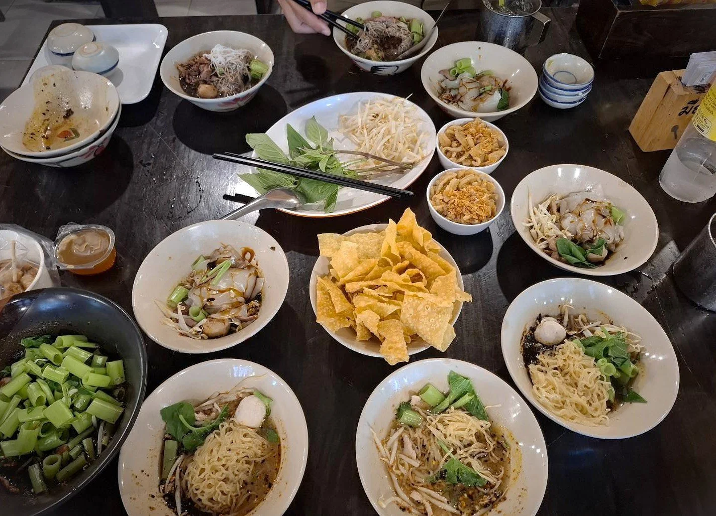 Table filled with assorted noodle bowls, crispy toppings, greens, and shared side dishes.