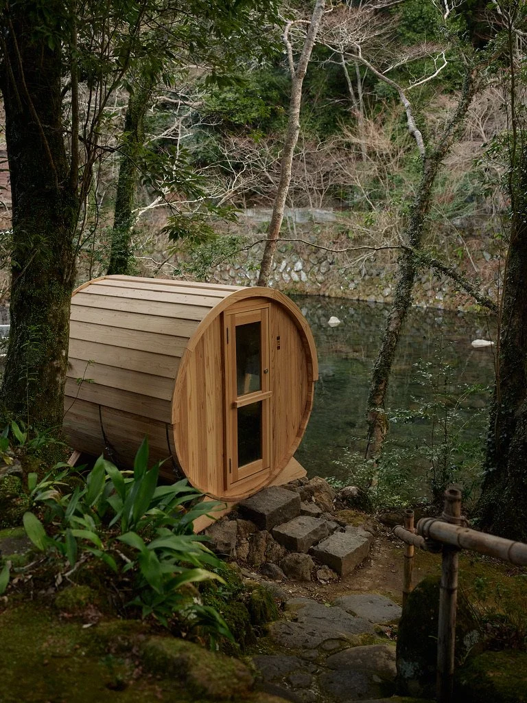 Small cylindrical wooden sauna with a rounded roof and glass door, set on a stone foundation beside water and surrounded by trees, rocks, and plants.