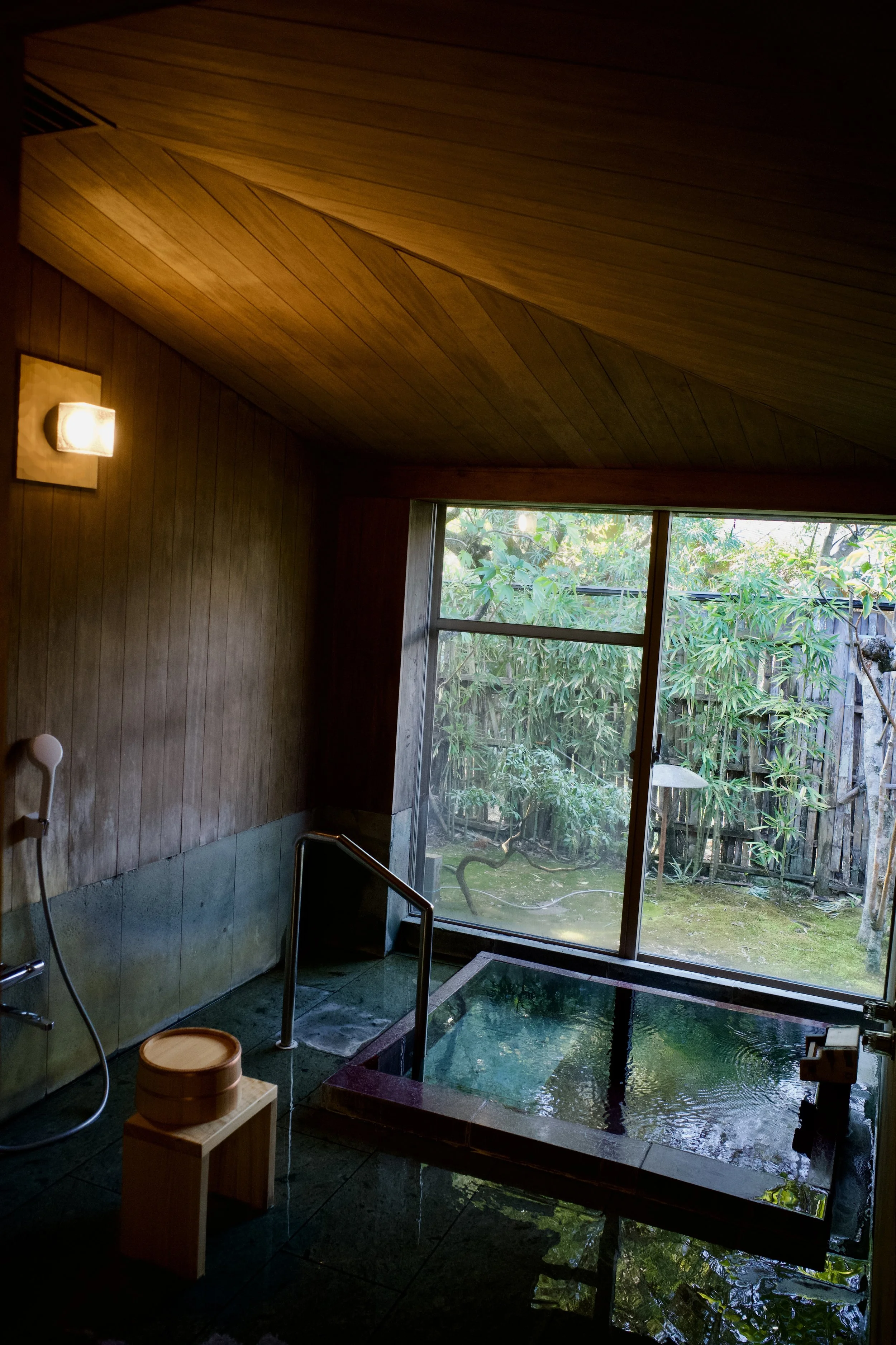 Indoor wooden bath with a rectangular soaking tub, a stool and bucket, and a glass door opening to a garden.