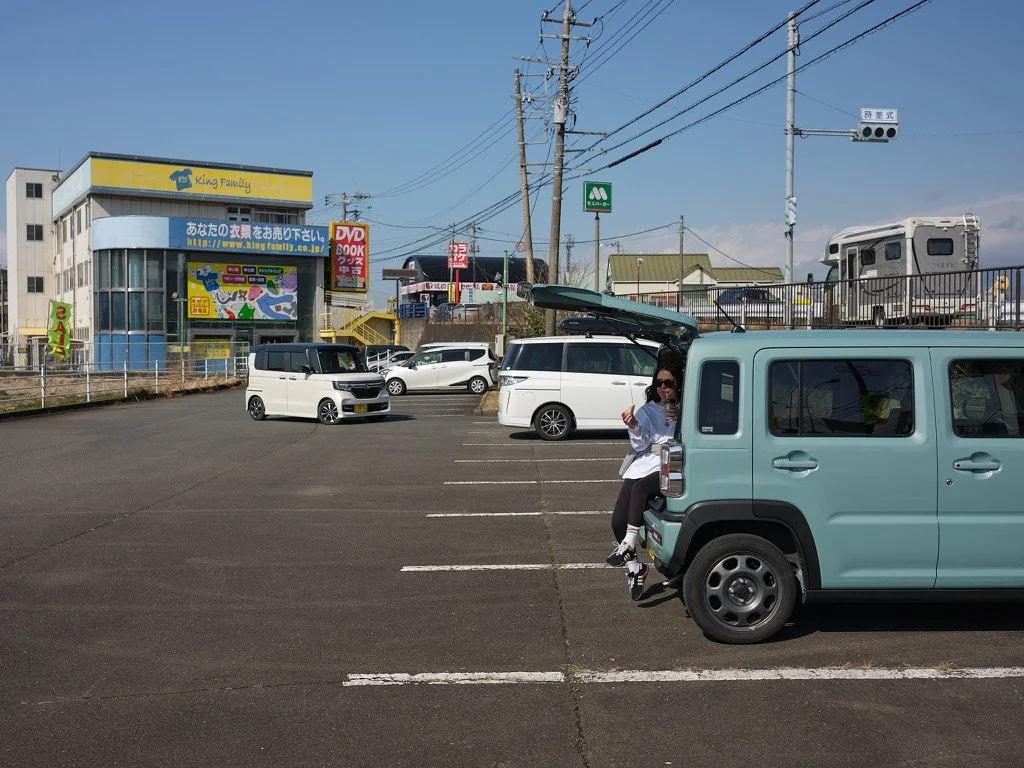 Parking lot in Izu with cars, a person, and stores in the background.