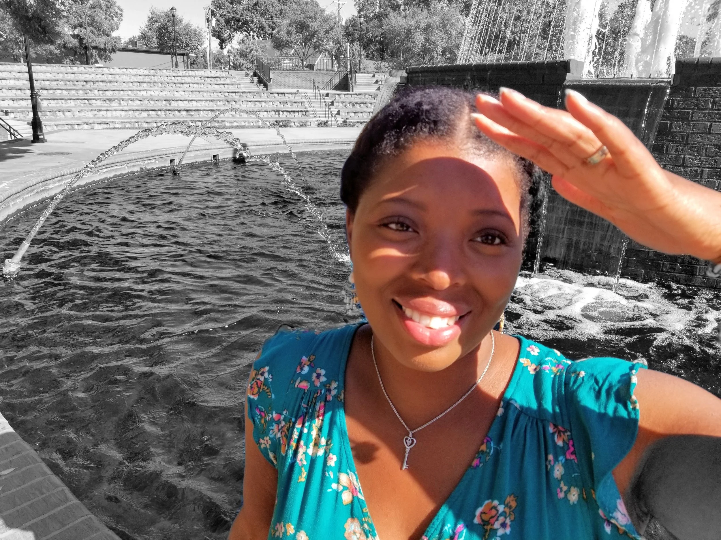 A smiling woman in a blue floral dress salutes with her right hand near her forehead, standing by a water fountain in a park, with steps and trees in the background.
