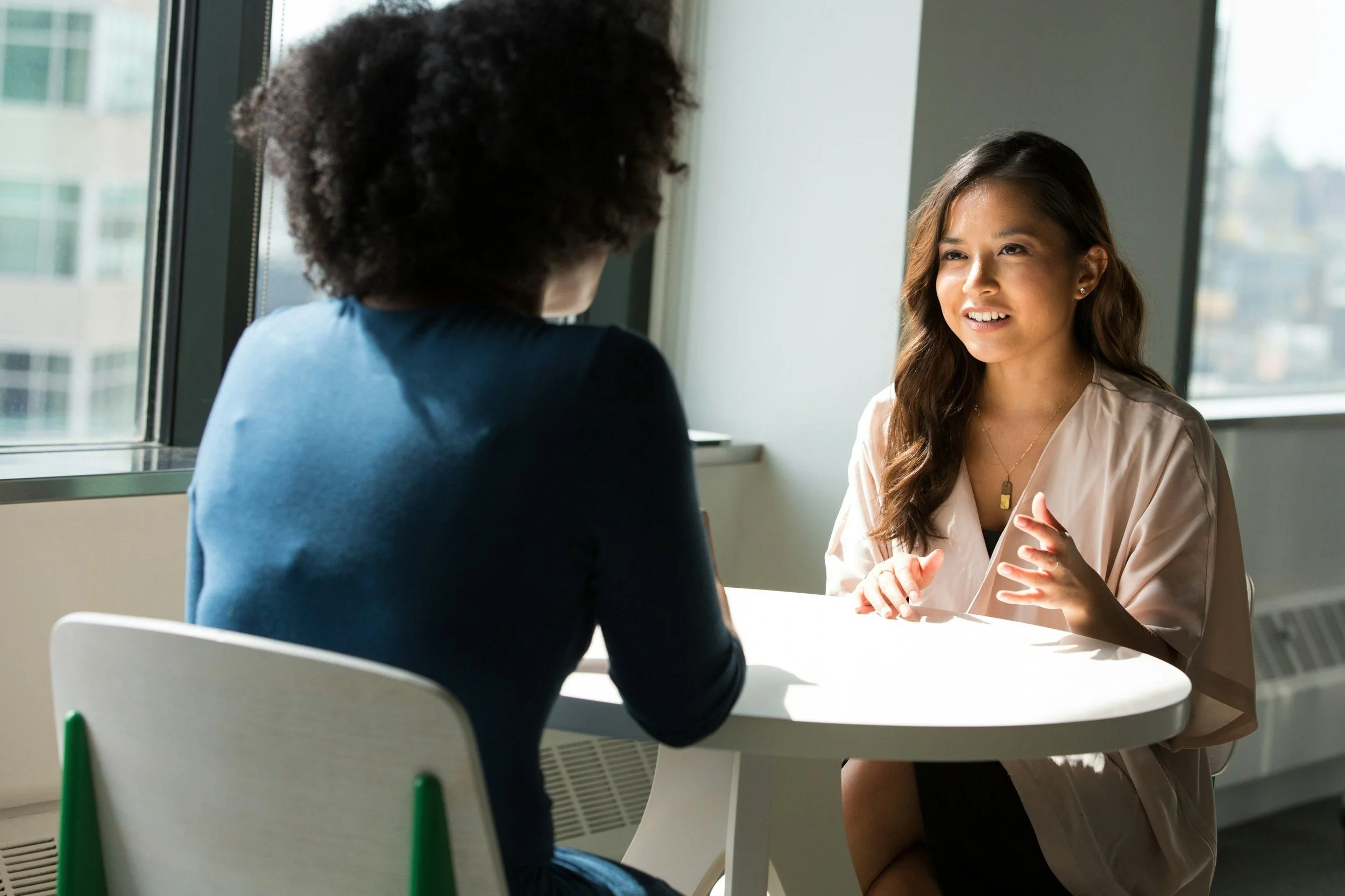Two women having a conversation at a round white table near large windows, with one woman speaking and the other listening.