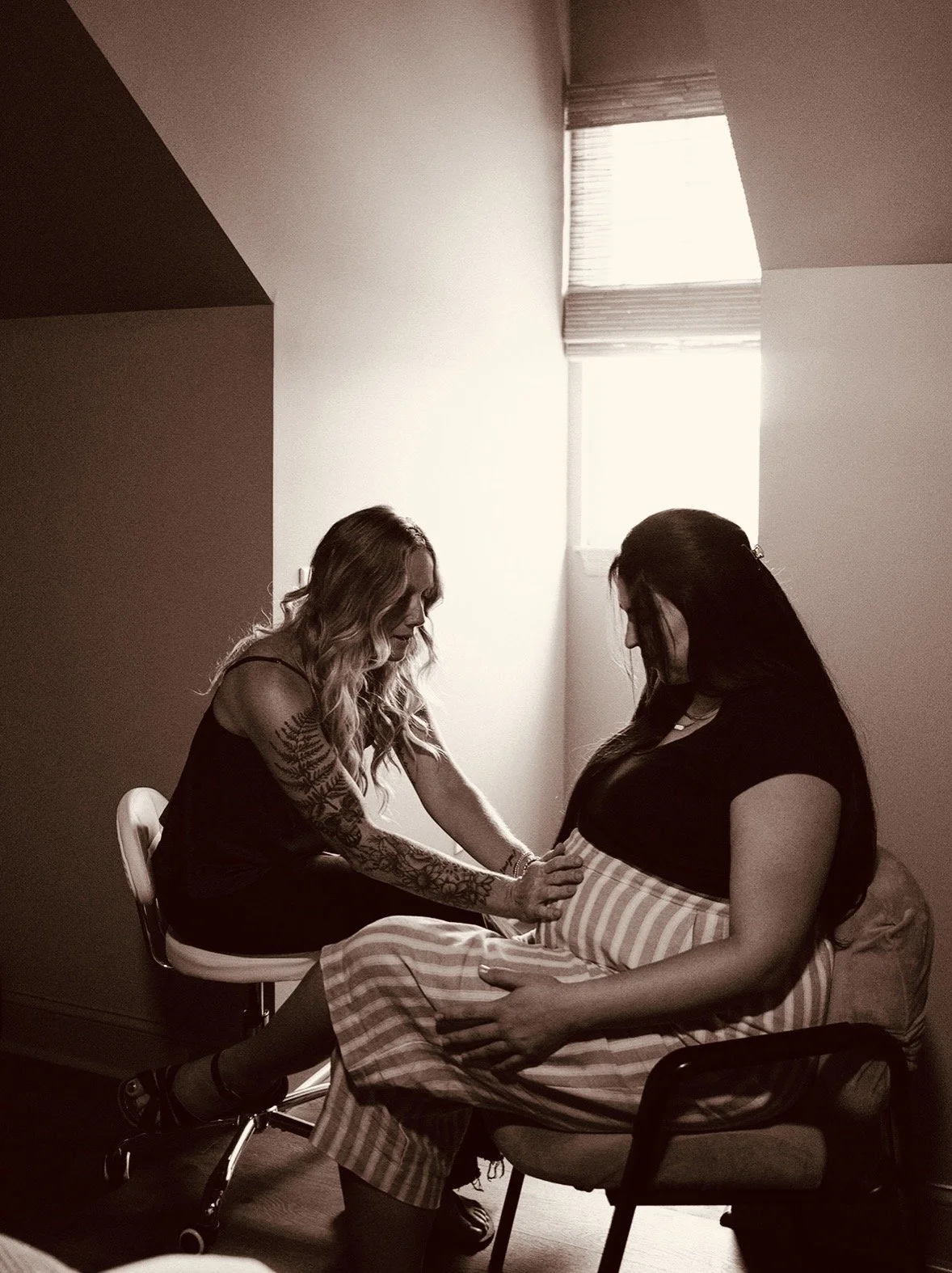 A woman in a hospital bed with a large pregnant belly, wearing a black bra, receiving support from an older woman leaning over and holding her shoulder in a hospital room.