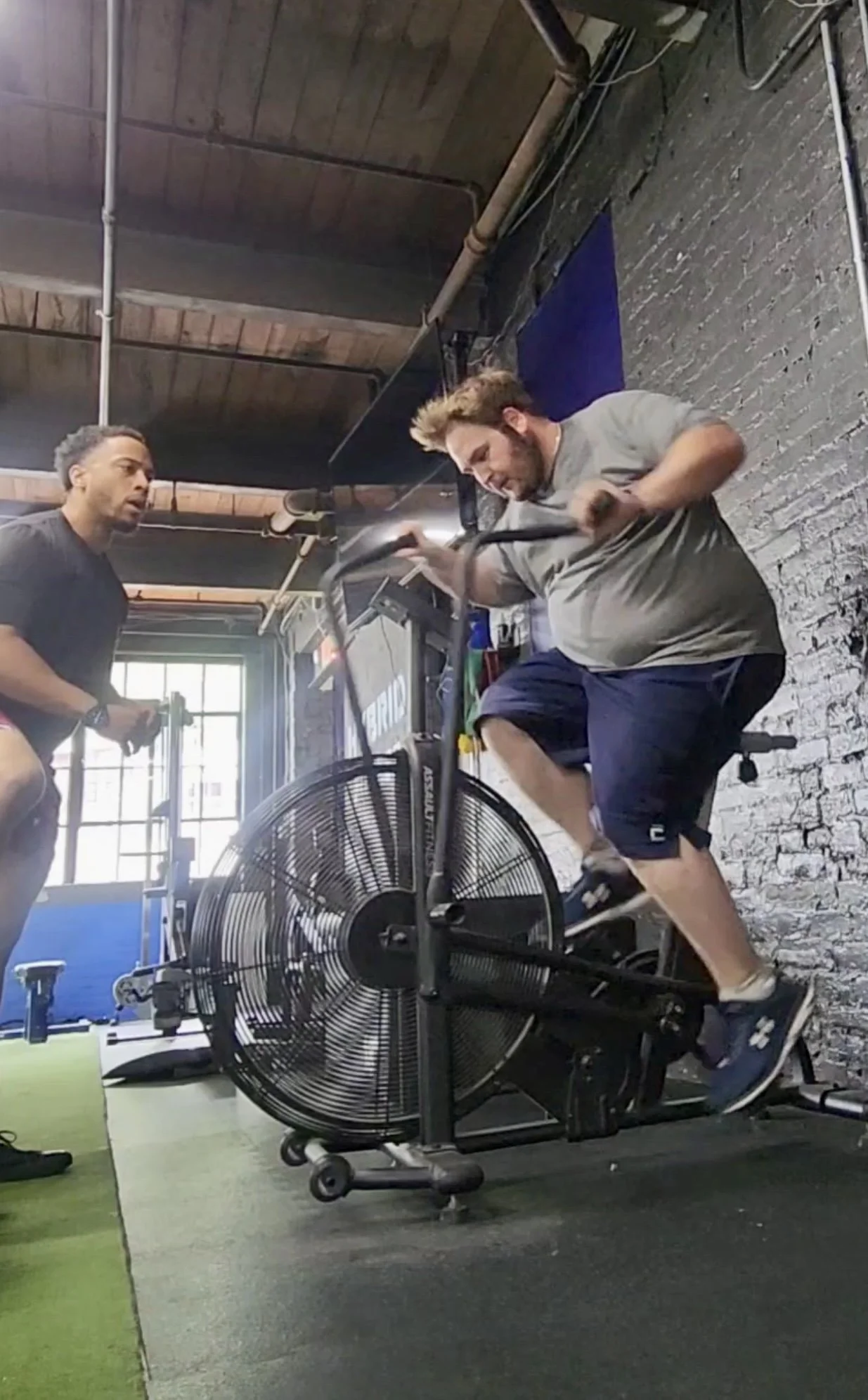 A man using an exercise bike while a trainer or coach observes in a gym with brick walls and high windows.