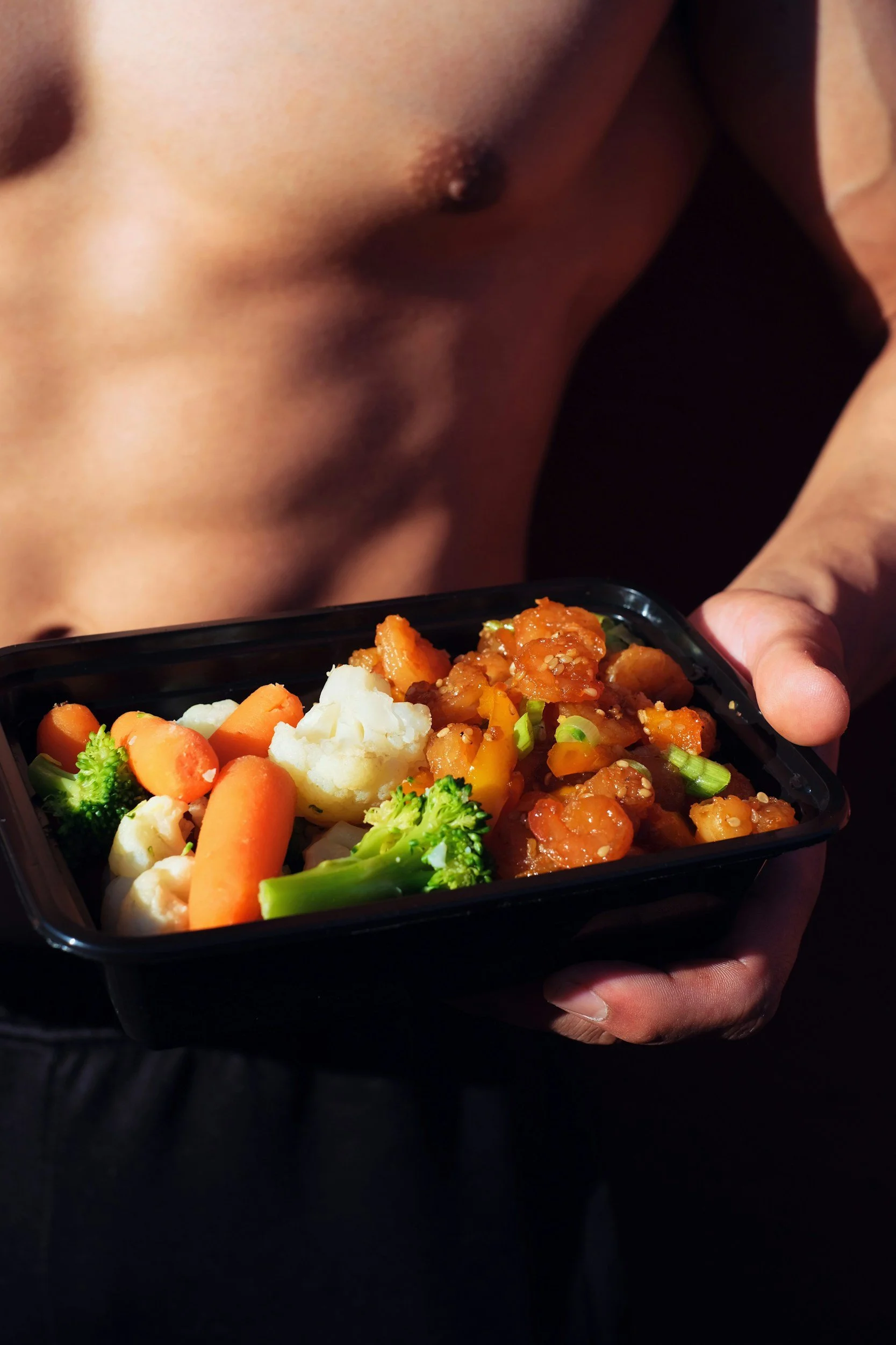 A person with a bare chest holding a meal container filled with mixed vegetables and a saucy dish.