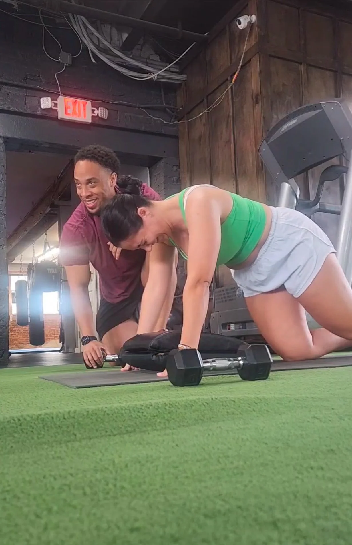 A woman doing a plank exercise on a mat with a dumbbell in front of her, smiling and looking down, in a gym with a man beside her and smiling.