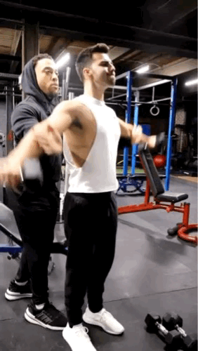 A man in a white sleeveless shirt lifting weights with assistance from a trainer behind him in a gym.