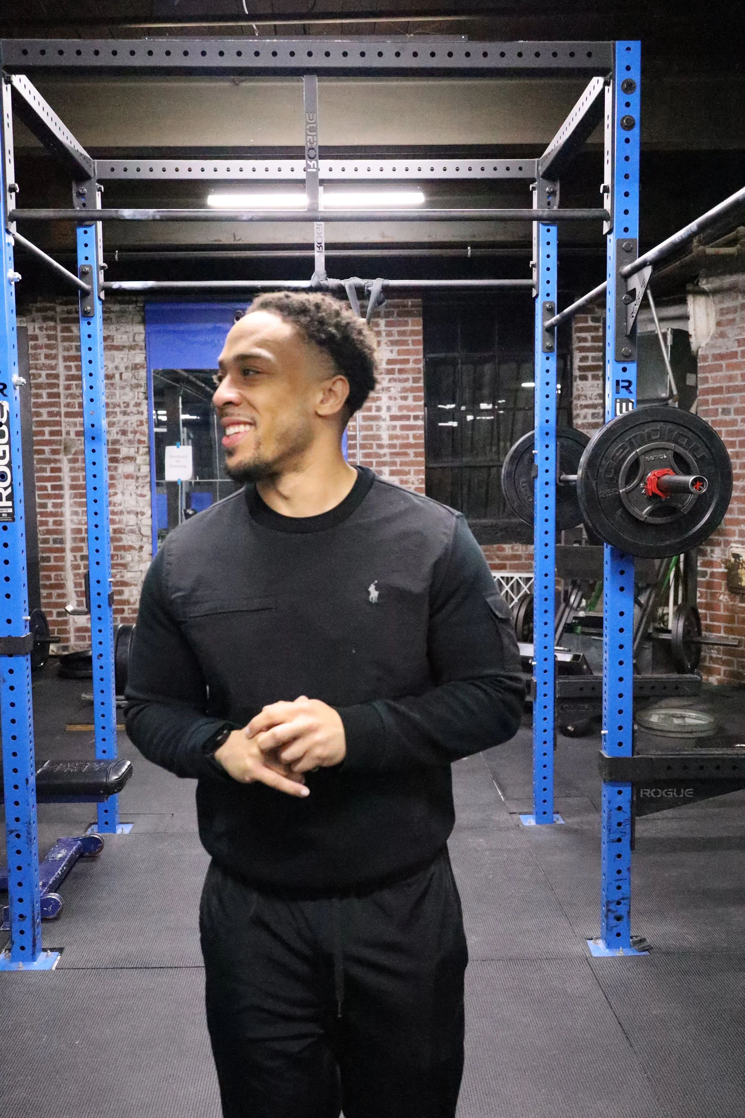 A man in a black long-sleeve shirt and black pants standing inside a gym in front of blue metal squat racks, with weight plates on the side and a brick wall in the background.