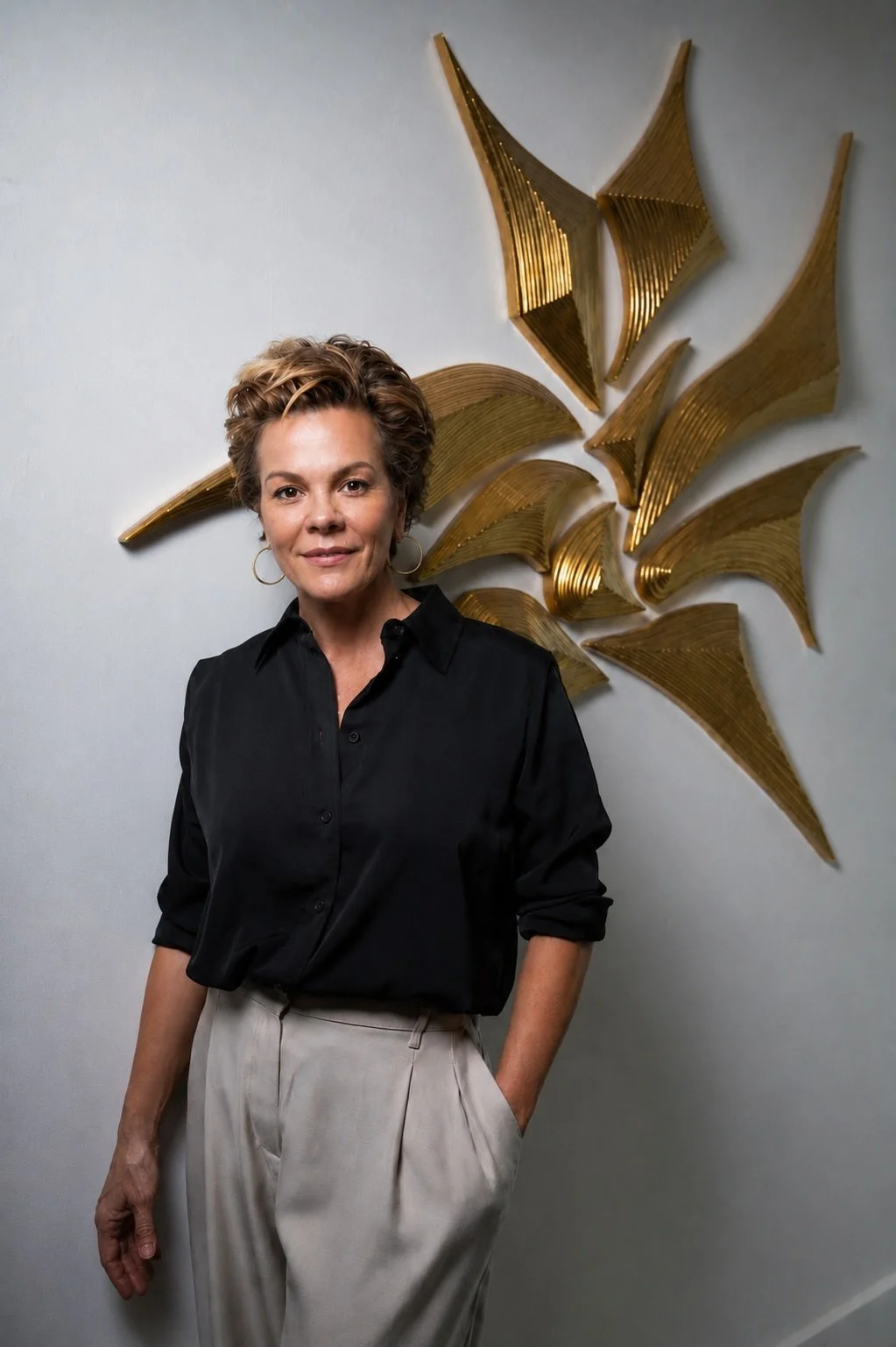 Jennifer Haley beside her gilded wall sculpture of layered, wing-like forms radiating across a pale wall, photographed at her Hollywood, Florida studio.