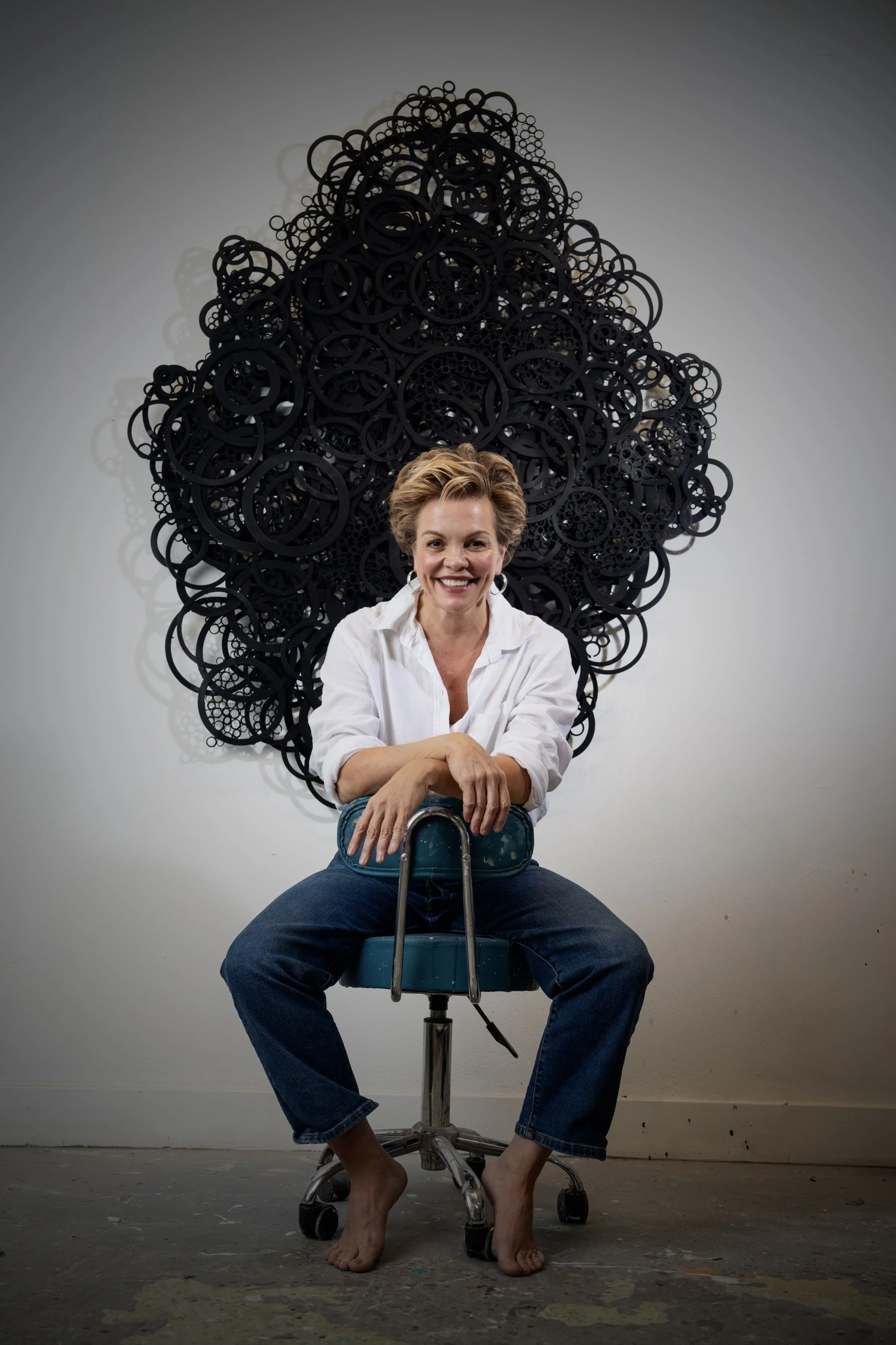 Jennifer Haley Studio abstract artist in front of her Algiz Rune inspaired wall sculpture called Algizian Veil made of matt black wall circles