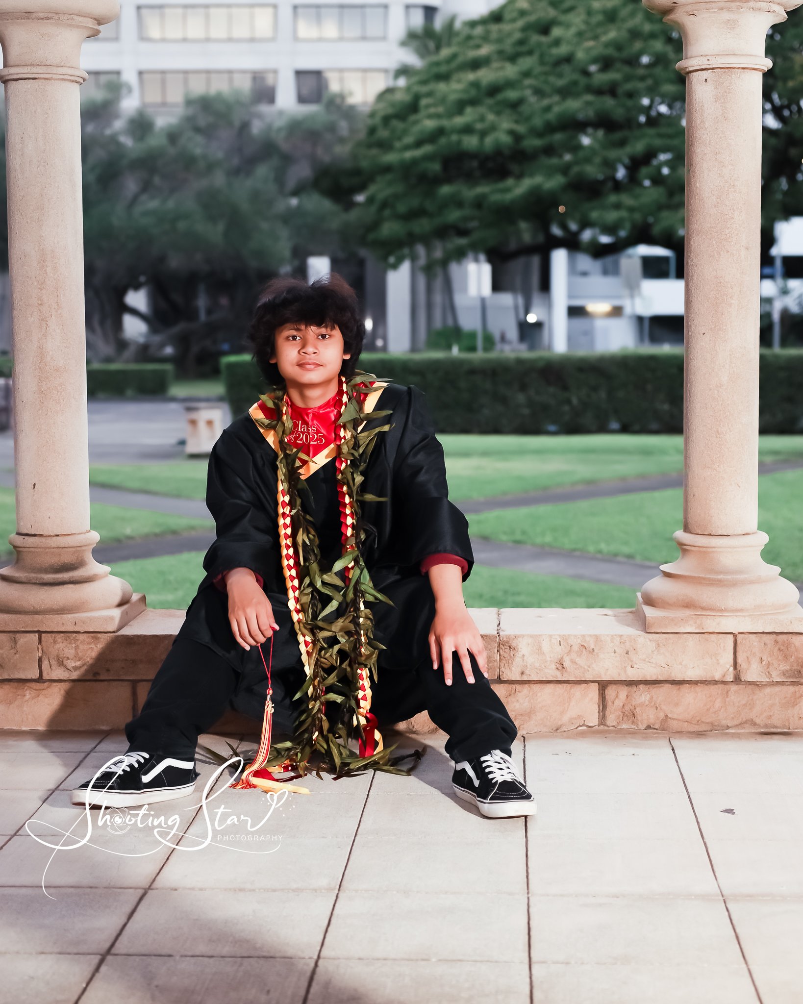 A young person sitting on a stone ledge in a park, wearing a graduation gown and cap, surrounded by greenery and urban buildings in the background.