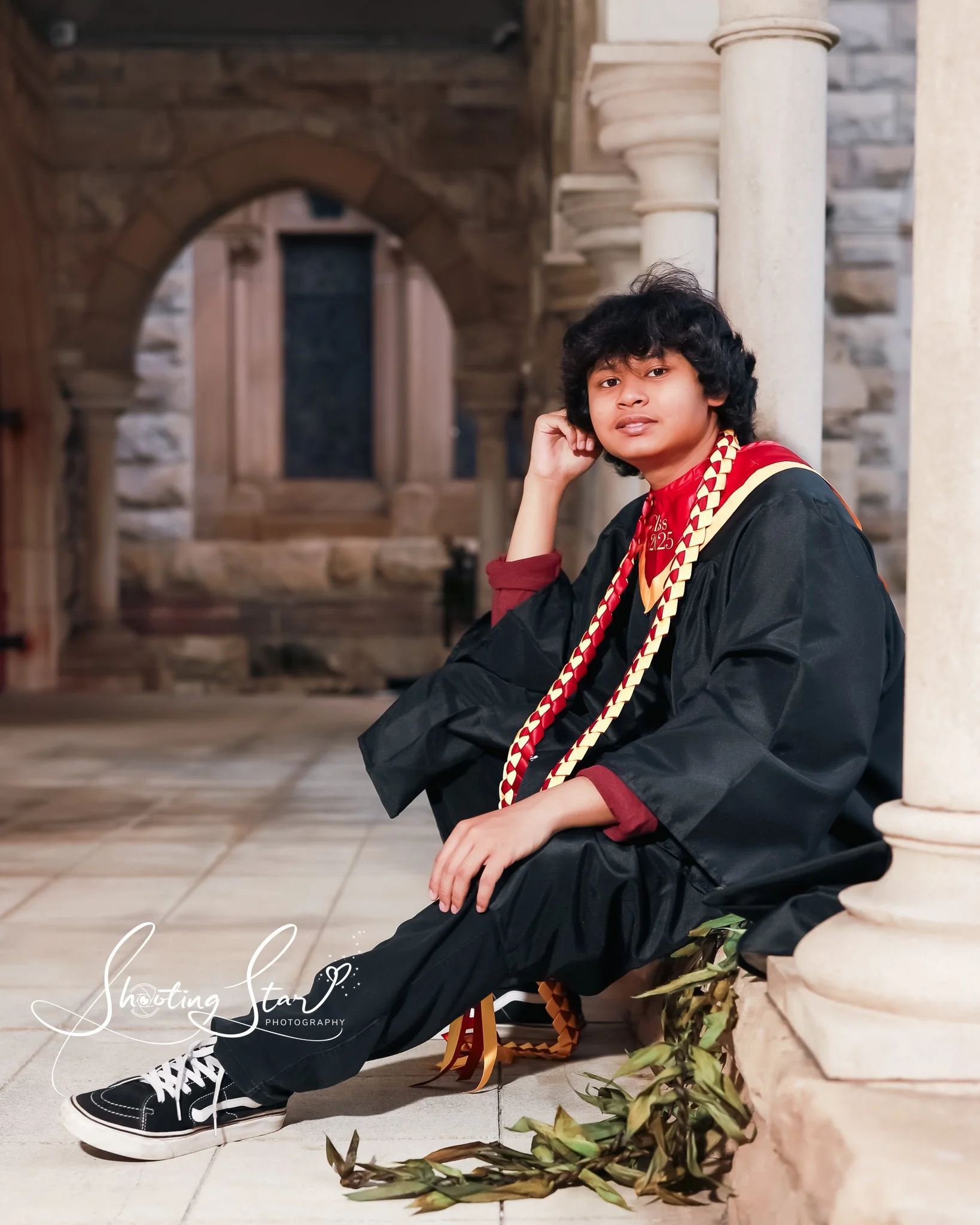 A young man with curly black hair sitting on the floor, leaning against a column in a stone and brick interior. He is wearing a black graduation gown, black sneakers with white laces, and a red shirt. He has a gold and red sash and a red and yellow c