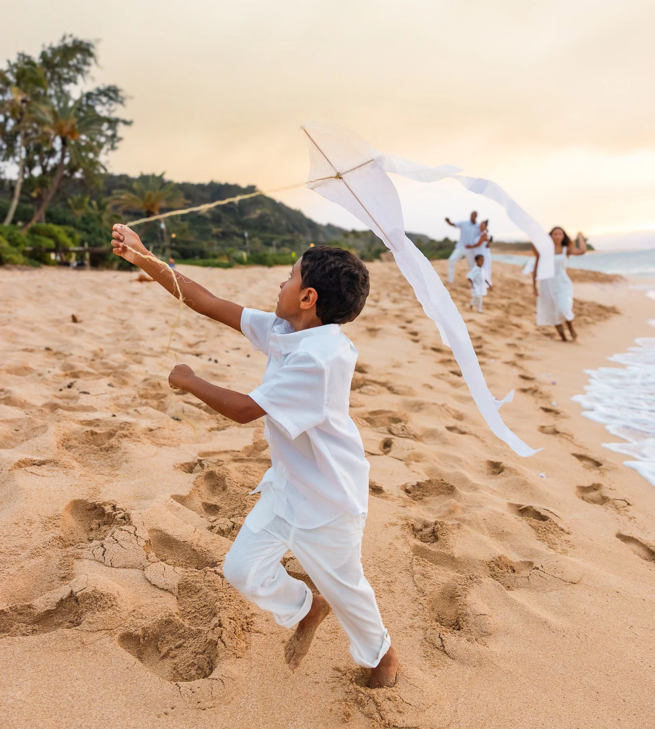 A boy in white clothing running on a sandy beach in Hawaii during sunset, flying a white kite with a long tail, with family in the background.
