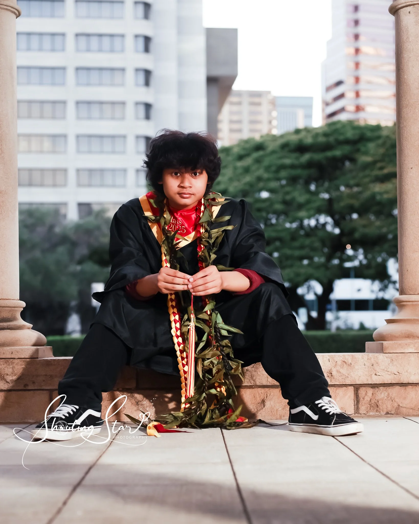 Young person in a graduation gown and cap sitting on a stone ledge outdoors, holding a green leafy garland with a red and gold ribbon, with tall buildings and trees in the background.