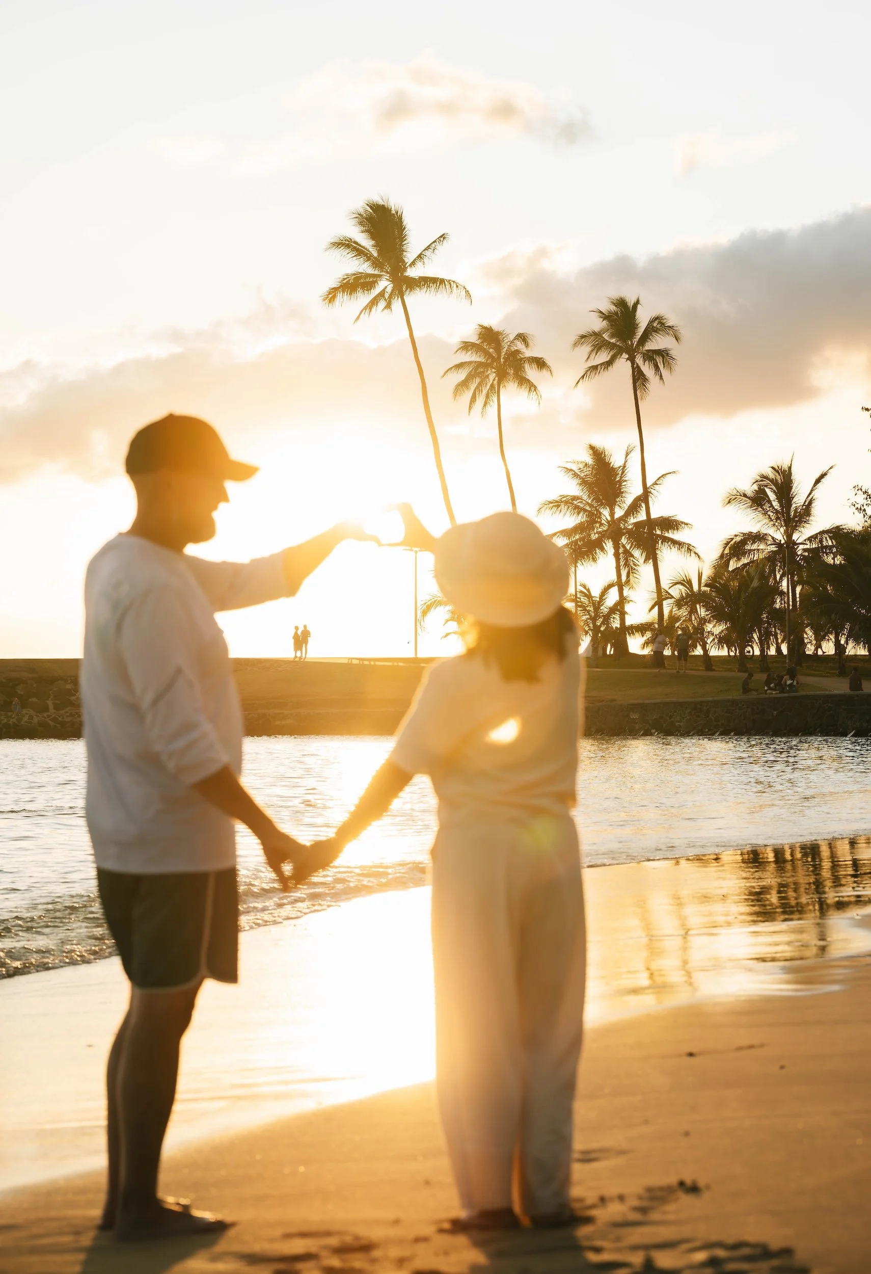 A couple holding hands on a sandy beach in Hawaii at sunset, with palm trees in the background and a person in the foreground wearing a hat.