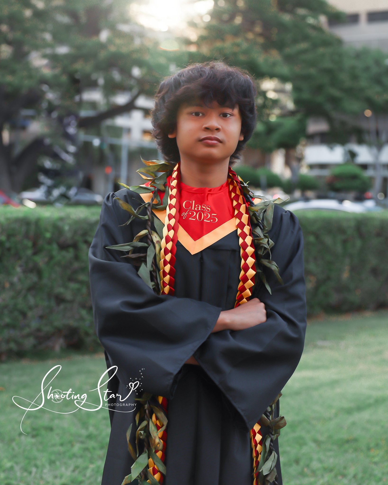 Young man in graduation cap and gown with leis, standing outdoors in a park or garden during daytime.