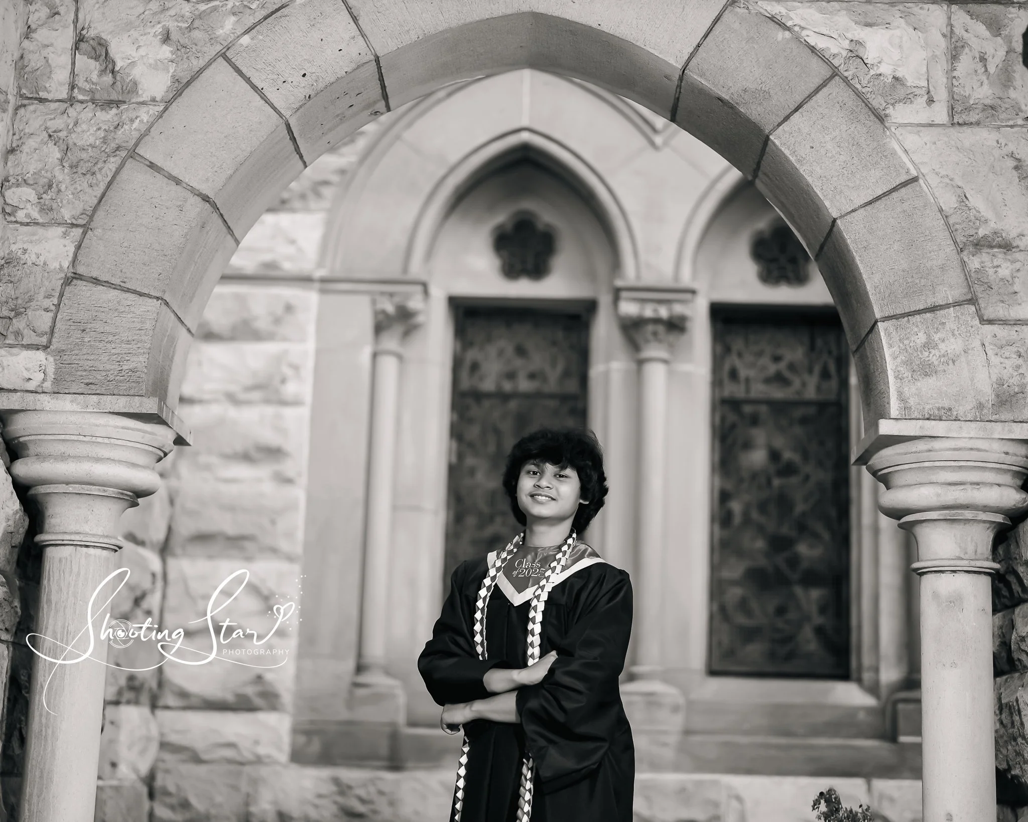 A young graduate in a cap and gown standing with arms crossed in front of a stone archway on a university campus, in black and white.