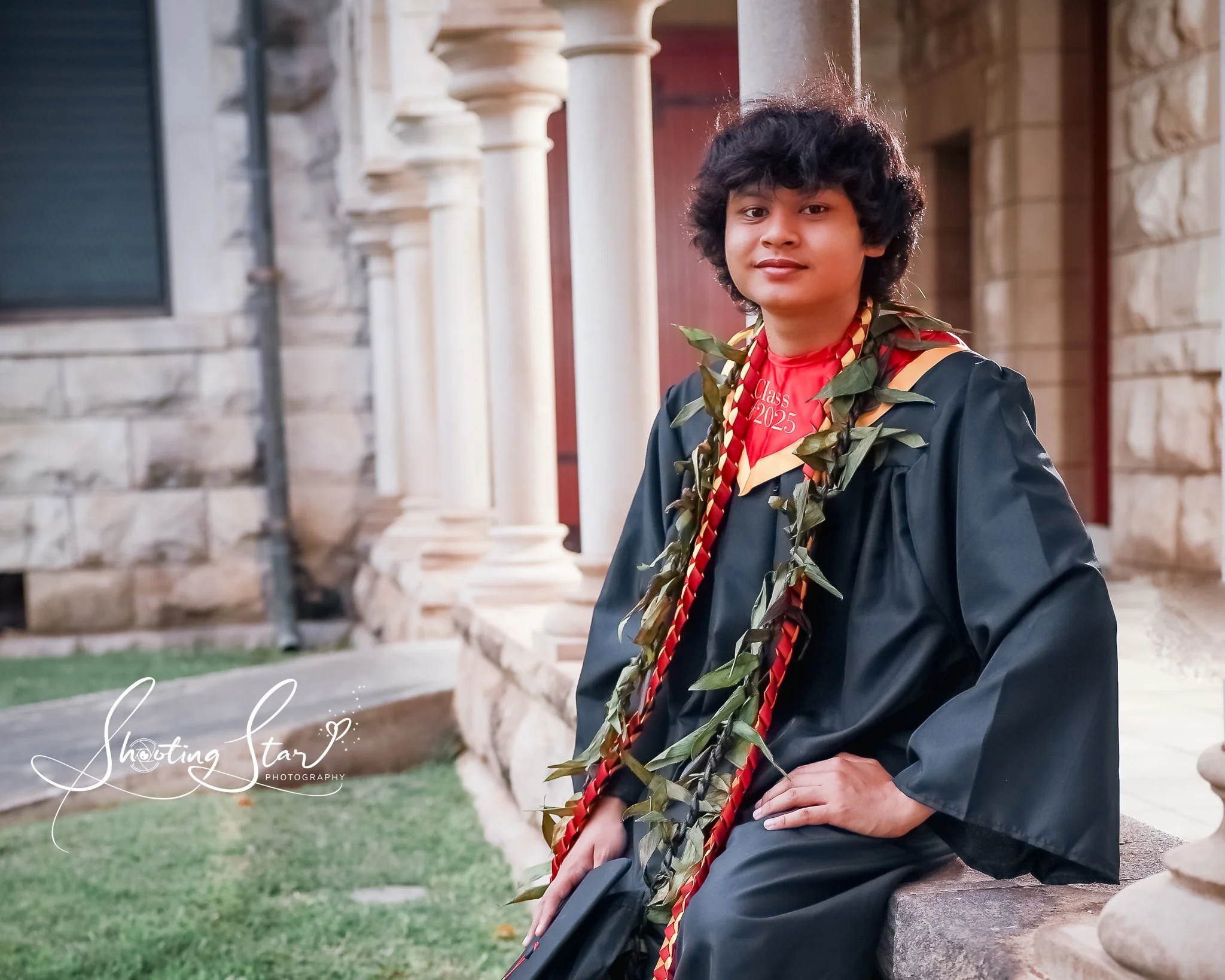 A young man in a graduation gown and medal, sitting outside on a stone bench, with a historic stone building in the background.