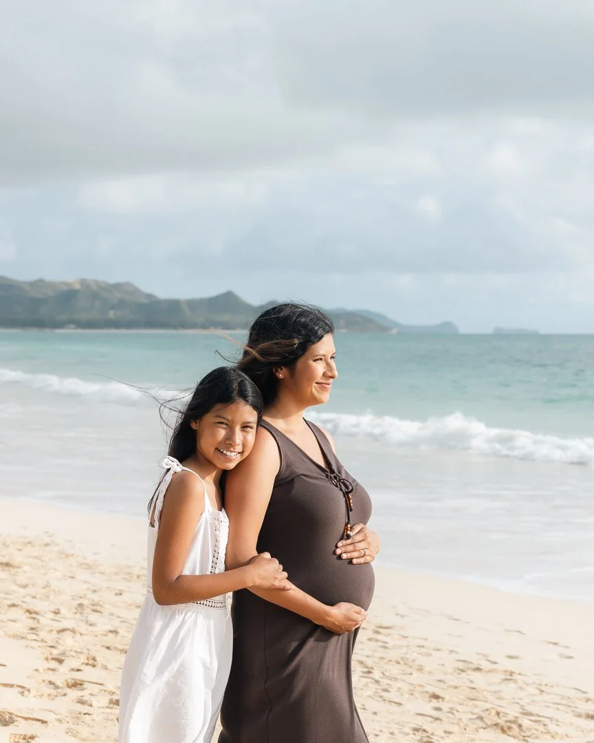 A mother and daughter on a beach, the mom is pregnant and wearing a brown sleeveless dress, while the girl is wearing a white dress. They are smiling and holding hands, enjoying the seaside scenery.
