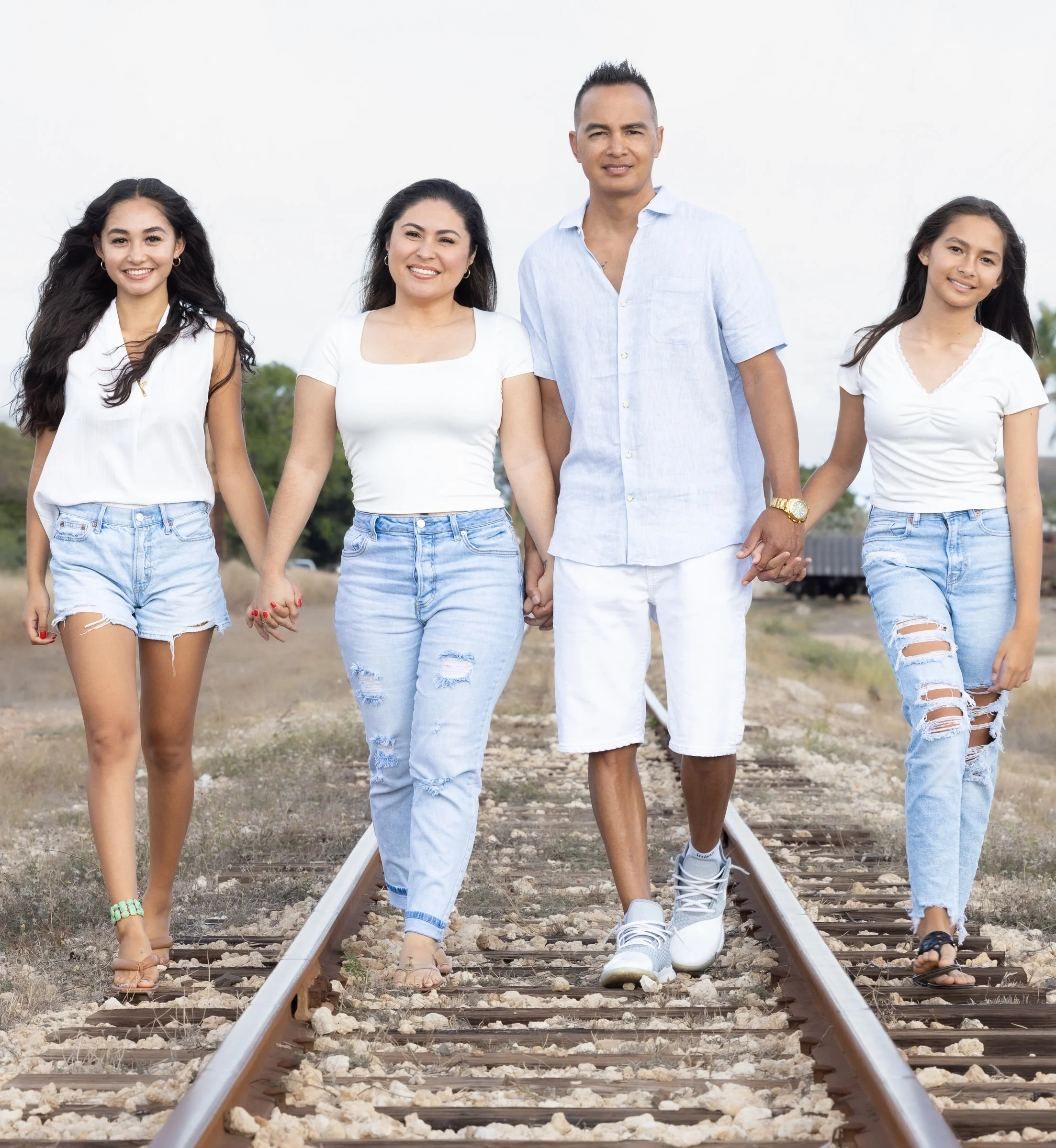 A mother, father and two daughters walking on a railway track outdoors with a green landscape in the background. They are smiling and dressed in casual white and light blue clothing, with three of them wearing sandals and one wearing sneakers.