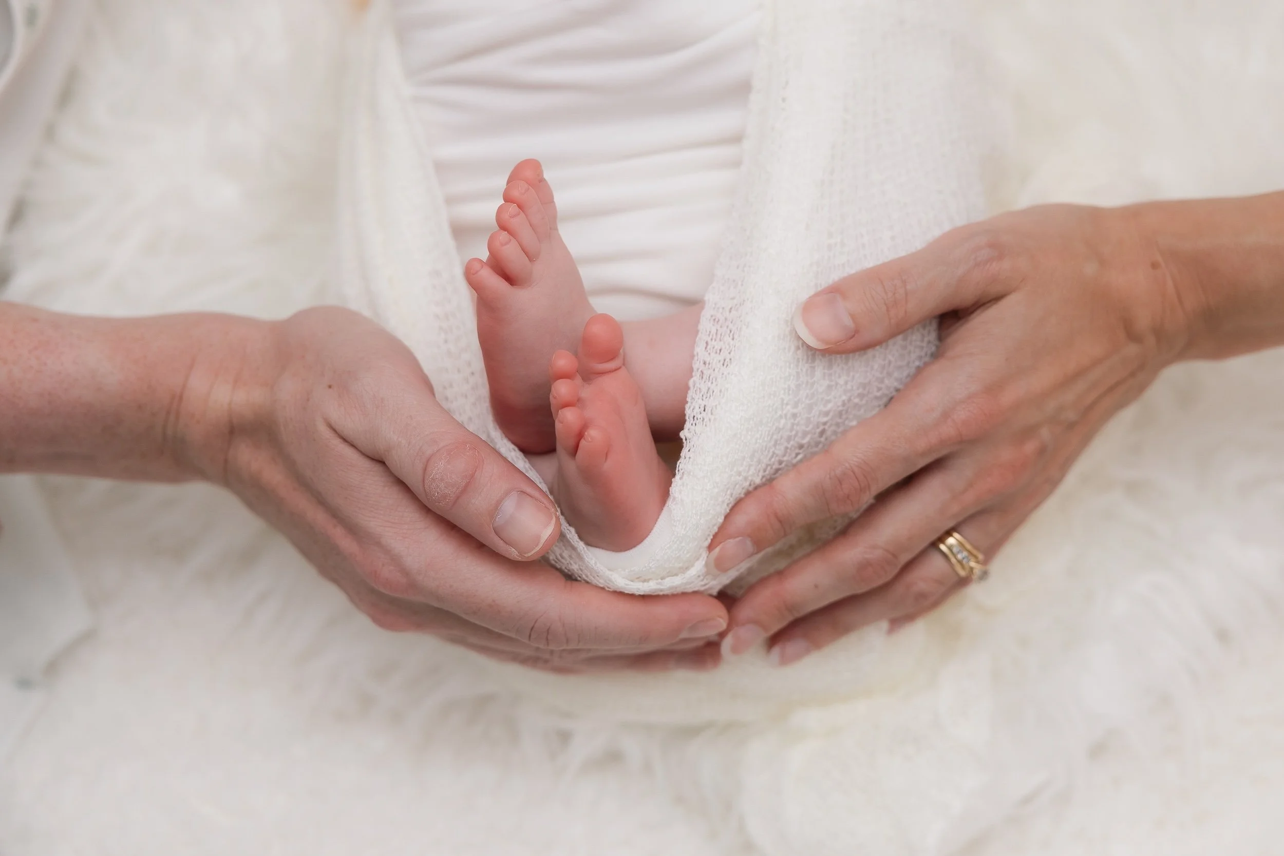 Close-up of a newborn baby’s feet held gently by an adult's hands, with the baby's tiny toes visible and wrapped in a soft, white blanket.