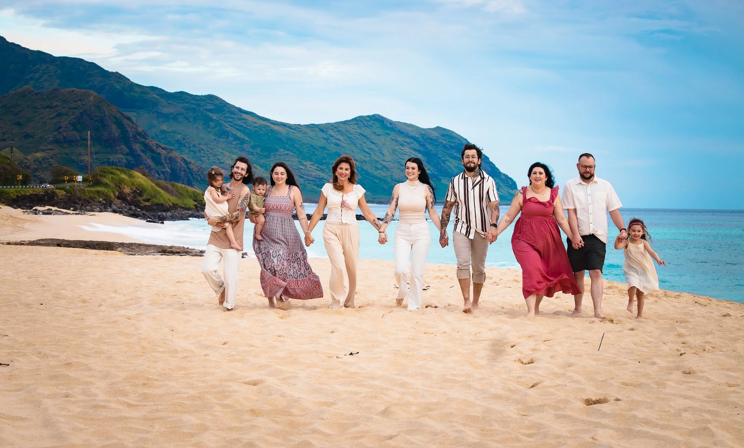 A family of eight people holding hands, walking on a sandy beach in Oahu with mountains and the ocean in the background.