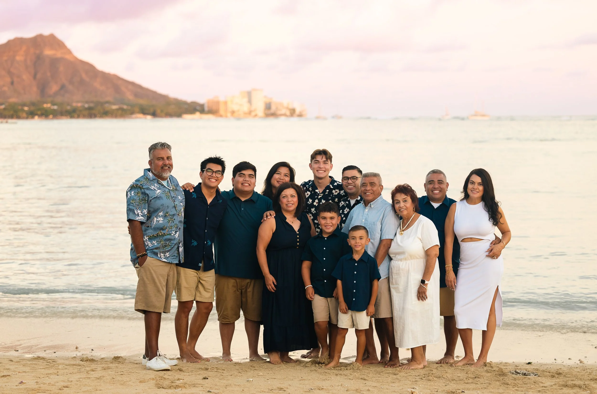 A family, including children and adults, standing on a beach in Oahu with water and mountains in the background, smiling at the camera.