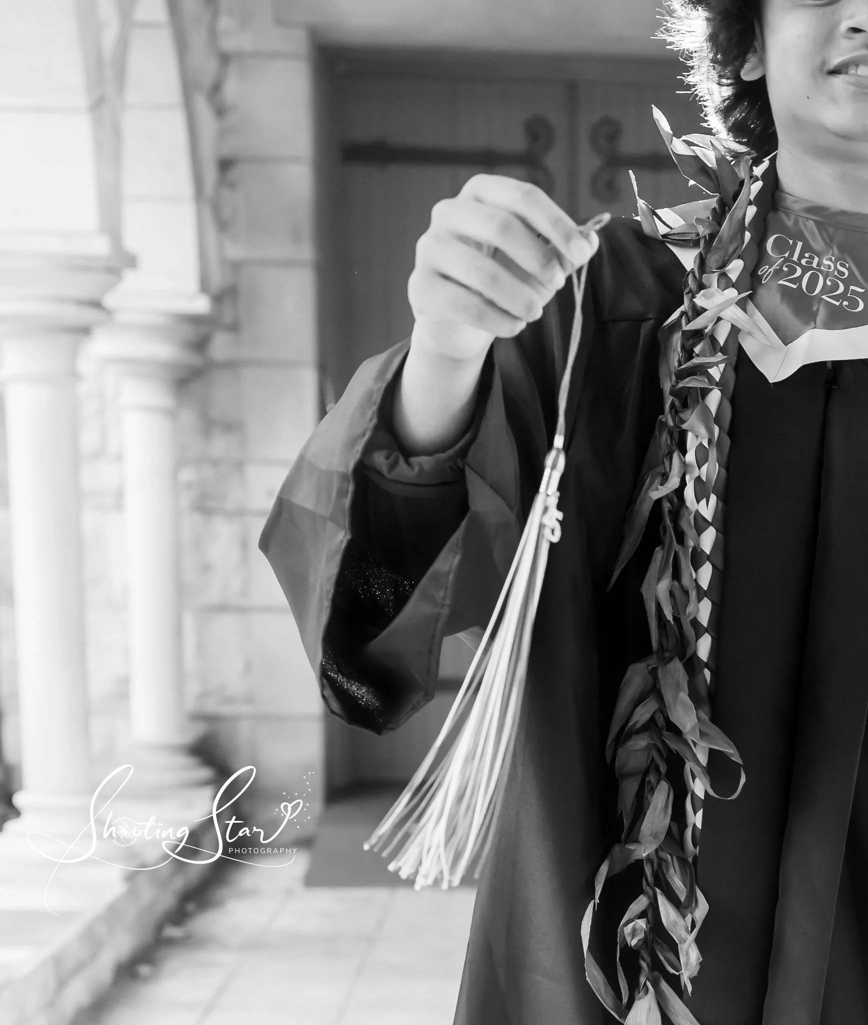 A graduation graduate holding a medal in their hand, wearing a cap and gown with a laurel lei around their neck, and a 'Class of 2025' collar pin.