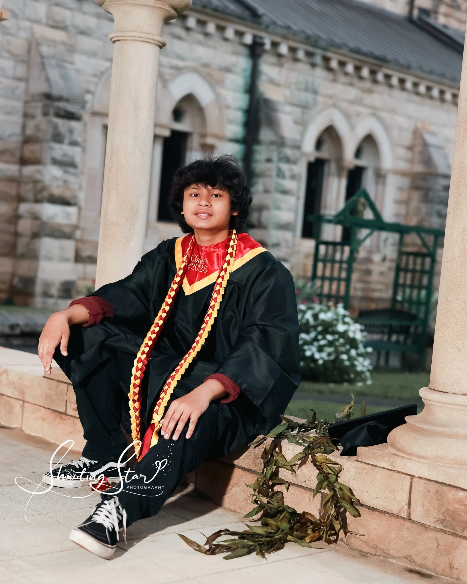 A young person wearing a graduation cap and gown, sitting on a stone ledge outside a stone building, with a diploma and a leafy branch nearby.