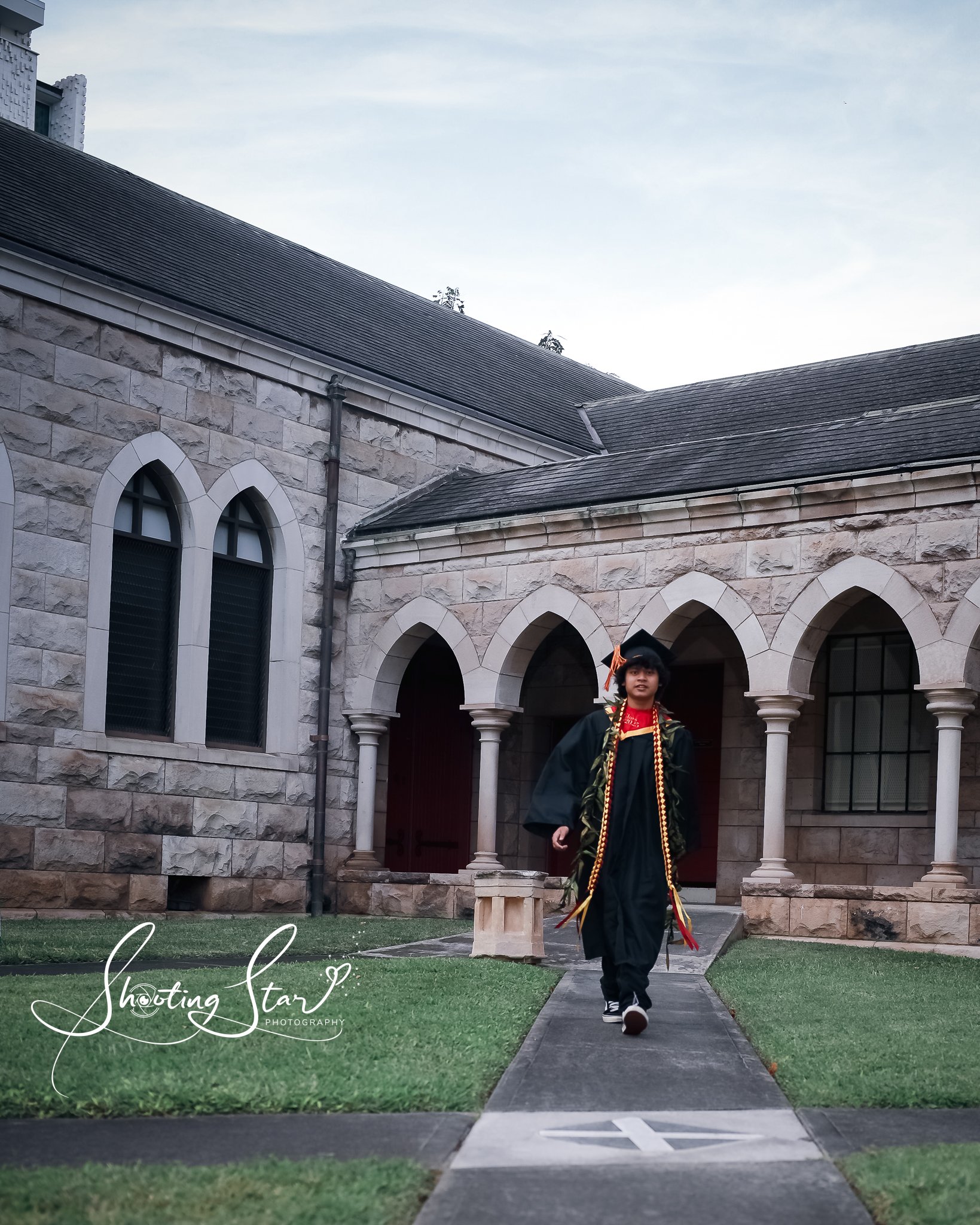 Person in graduation gown and cap walking on a pathway outside a stone building with arched windows and columns.