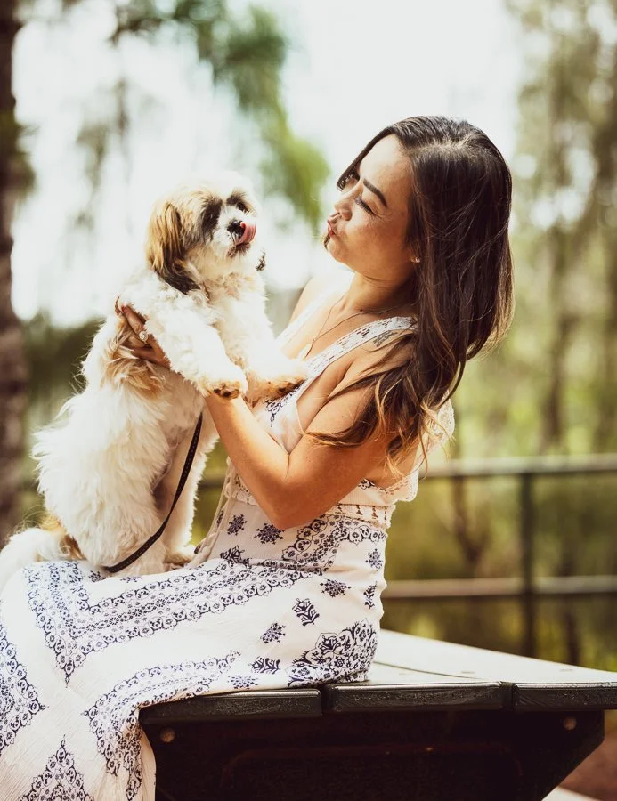 A woman sitting on a bench outdoors holding a small fluffy dog, appearing to kiss the dog.