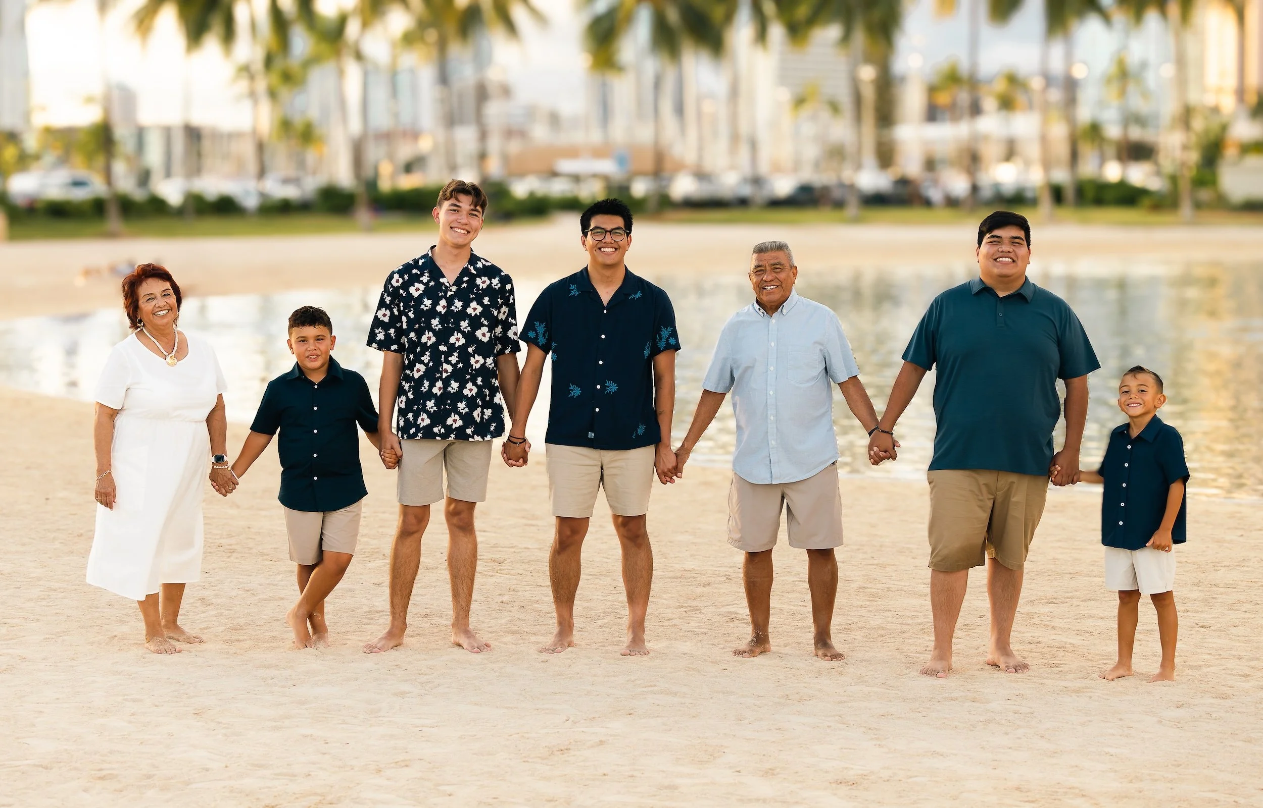 Multi-generational family holding hands and walking on the beach, smiling, with a city in Hawaii in the background.
