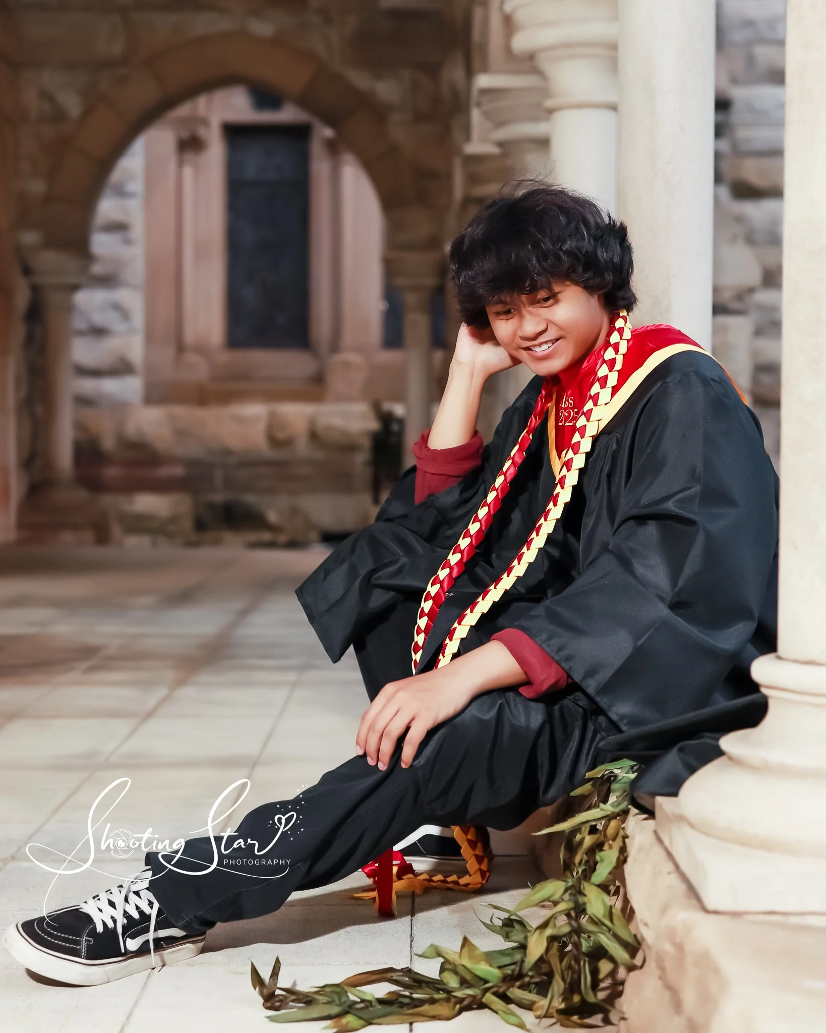 A young person sitting on the ground in a graduation gown and sandals, smiling and looking down, with leaves on the floor, indoors with stone walls and columns in the background.