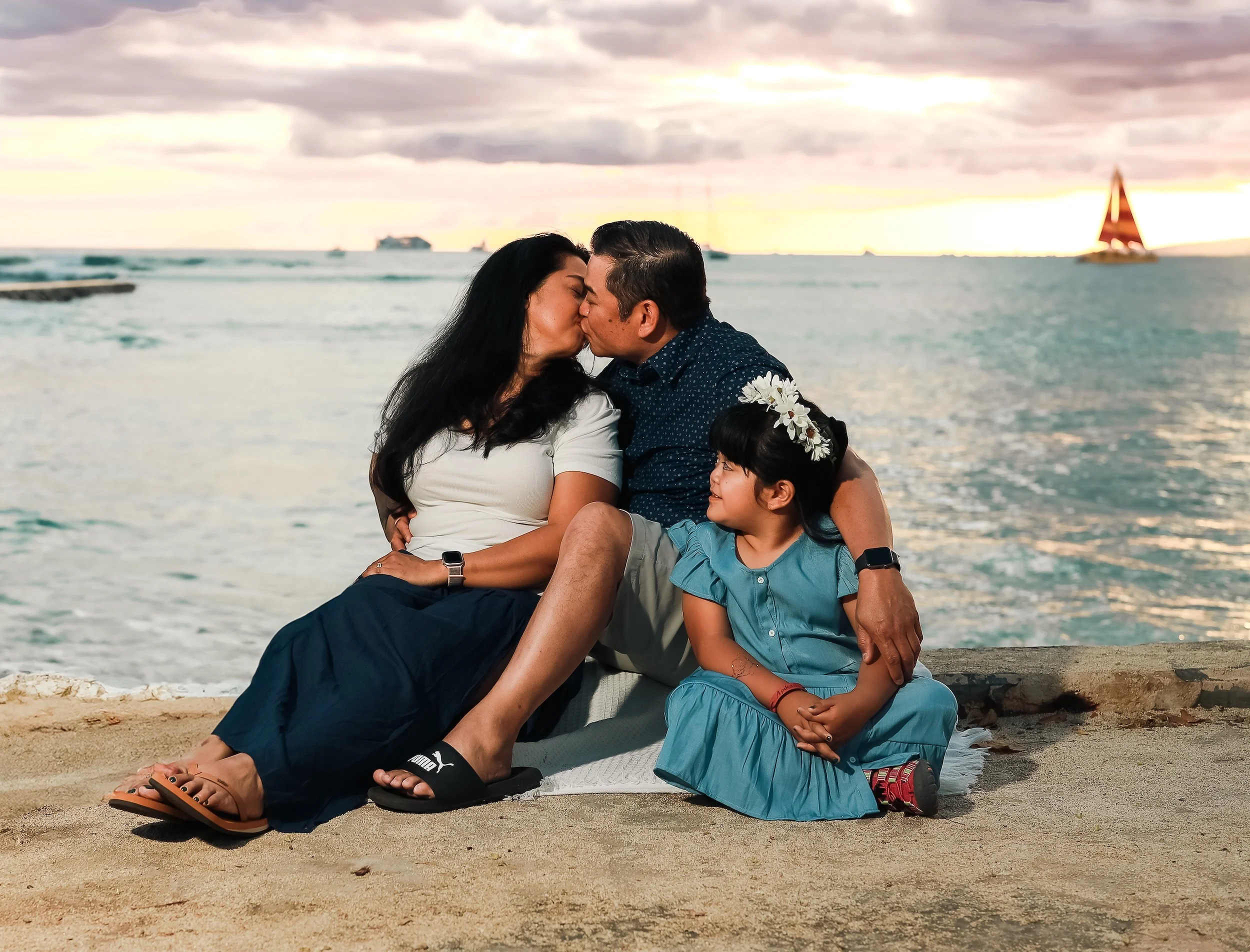 A family of four sitting on the sand at the beach, sharing a kiss with the ocean and boats in the background during sunset.