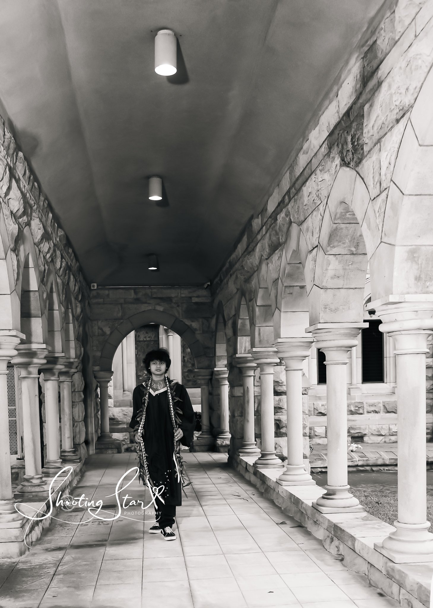 Black and white photo of a person in a graduation gown and cap standing in an arched marble corridor with columns.