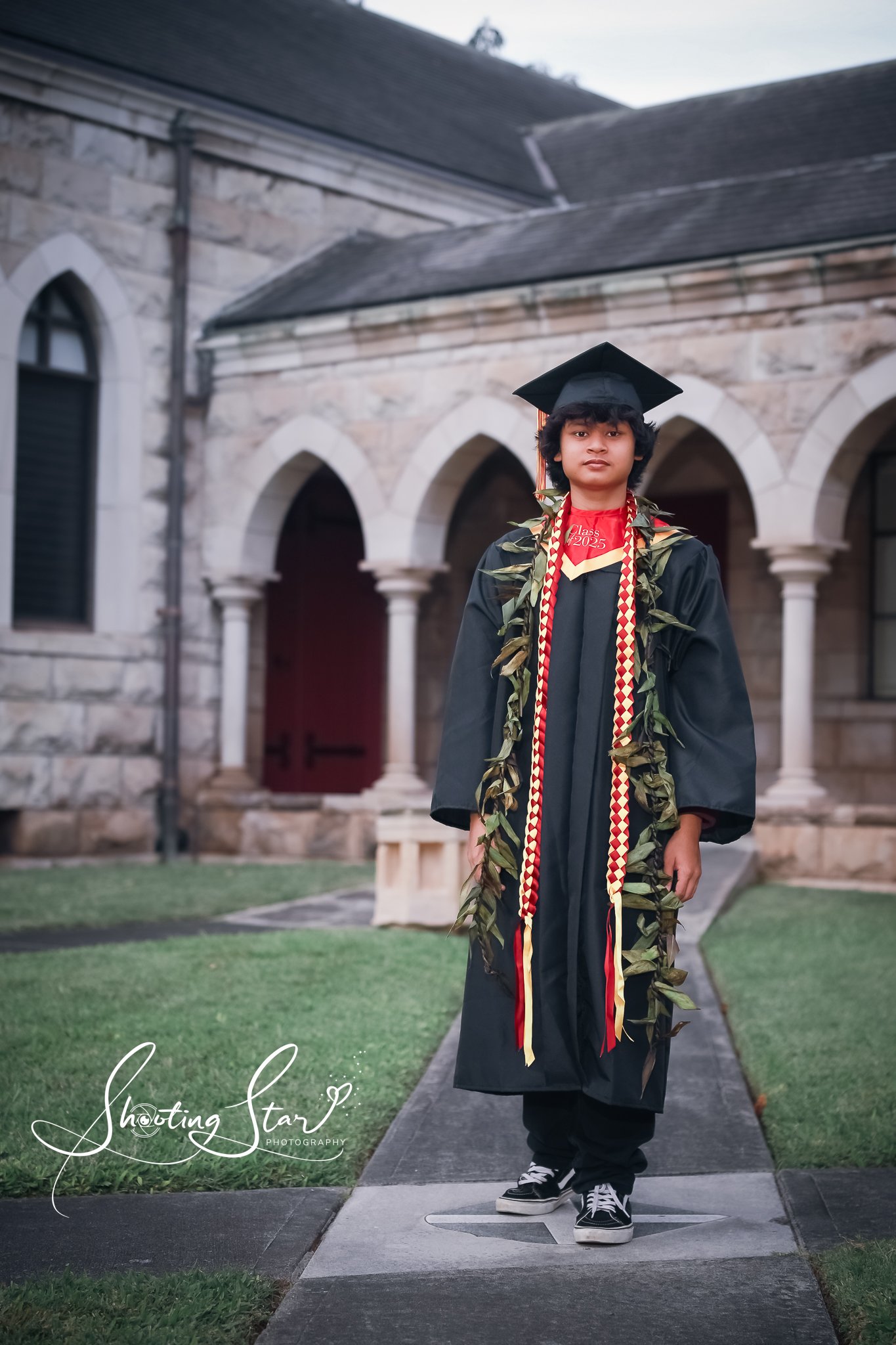 A young graduate in cap and gown standing outdoors on a pathway in front of a stone building with arched windows and columns.