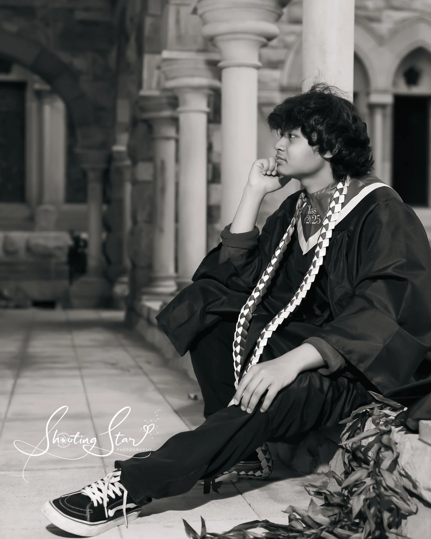 A young man in graduation attire sitting on the ground, resting his chin on his hand, with a thoughtful expression. He is wearing a black robe, a graduation stole, and Vans sneakers, and is seated near columns and European-style architecture.