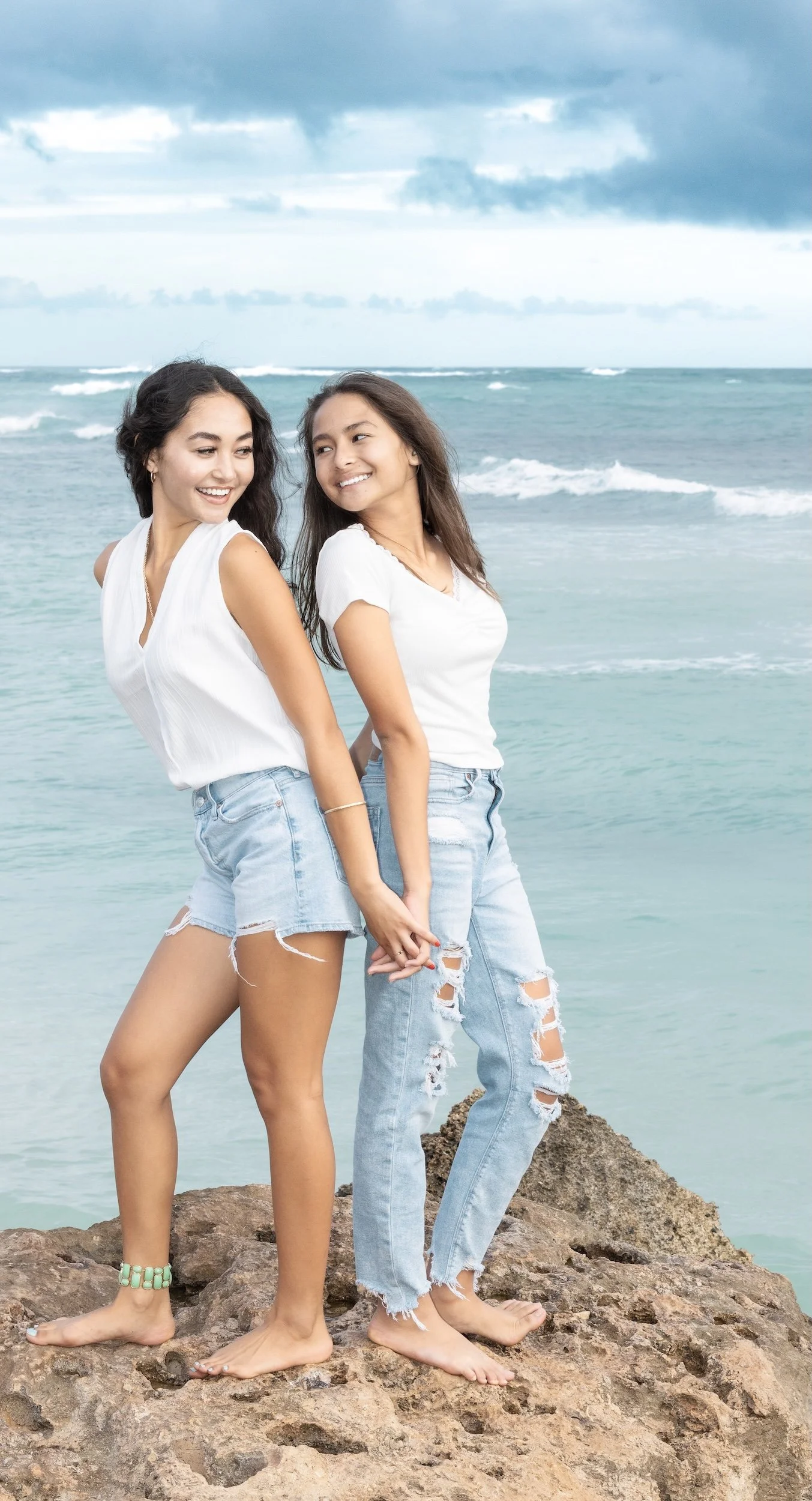 Two young sisters in white t-shirts and ripped jeans standing on rocks near the ocean, holding hands and smiling at each other with a cloudy sky in the background.