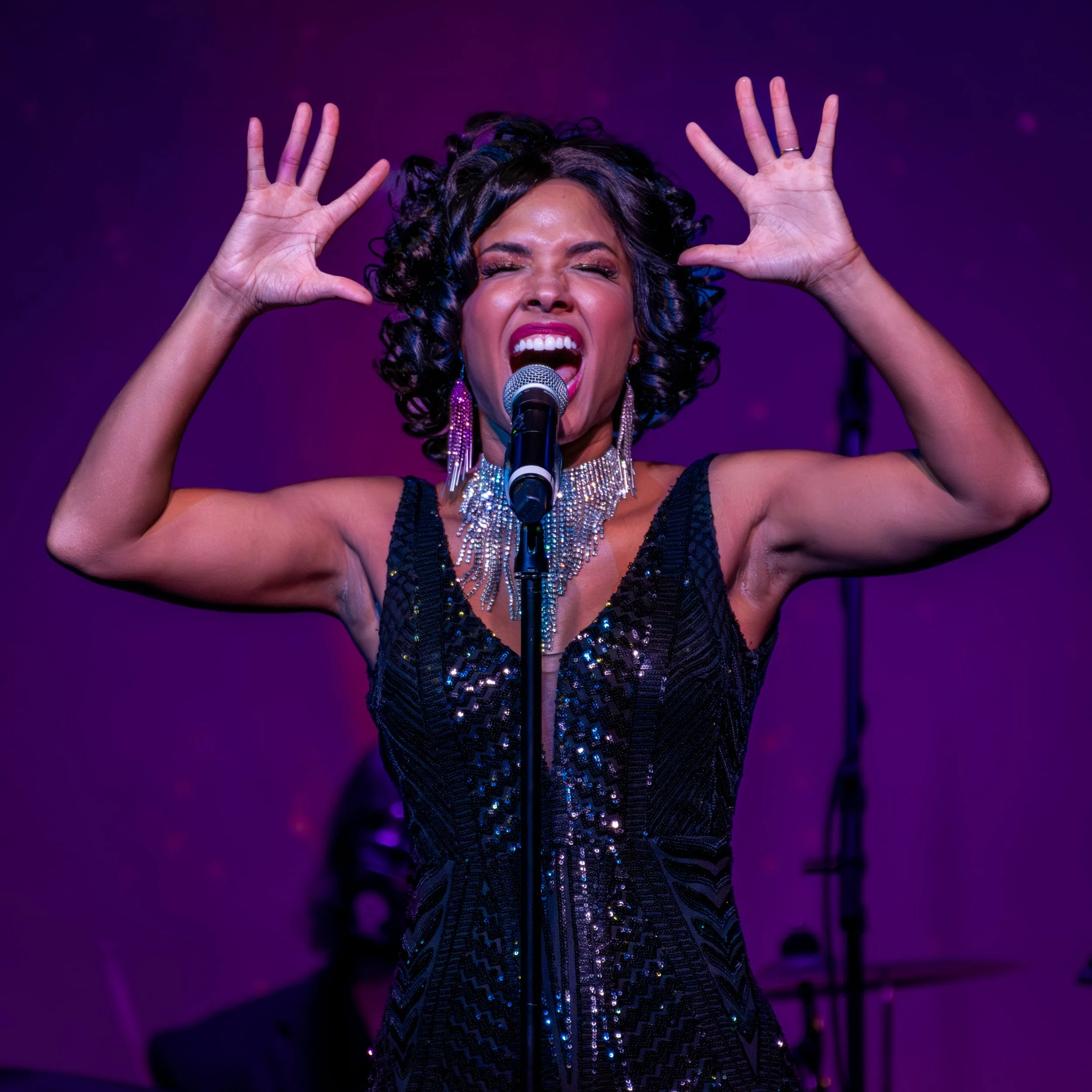 A woman with curly dark hair singing passionately into a microphone on stage, with her arms raised and hands forming a gesture, wearing a sparkling dress and earrings, against a purple background.